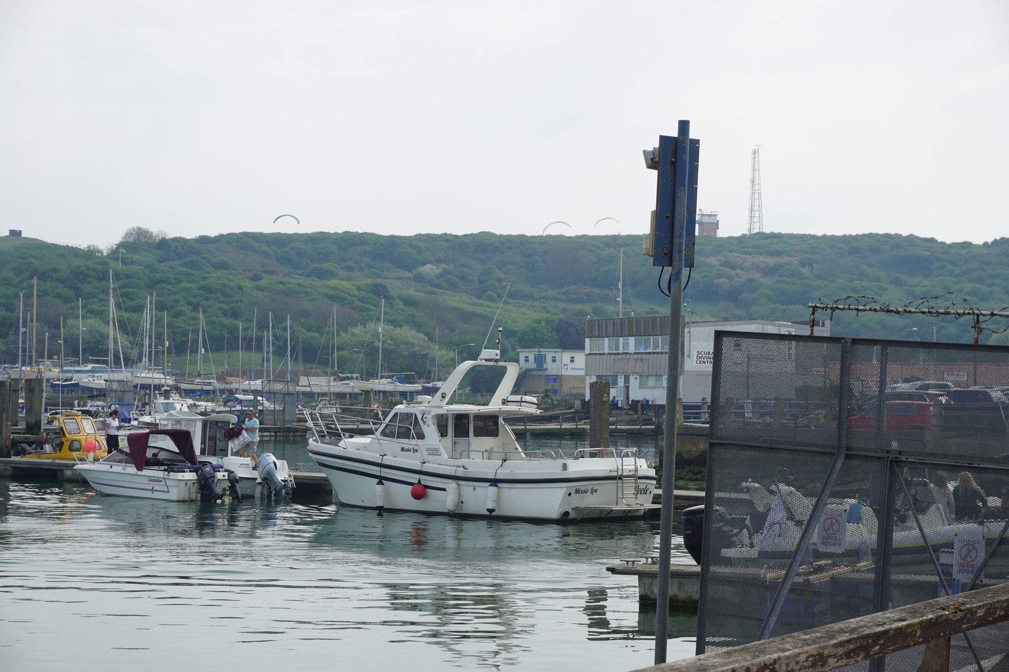 Boats in a marina. In the distance, some large kites can be seen.