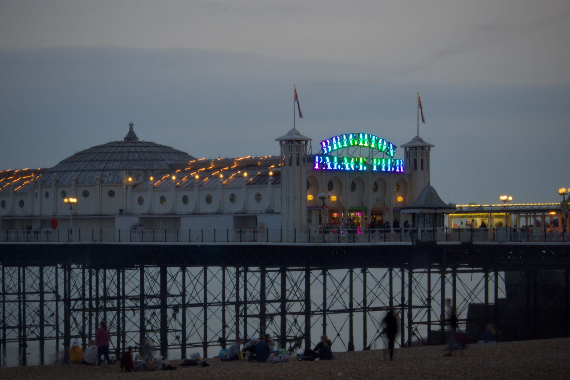 Brighton Pier at dusk. People walk along the beach.