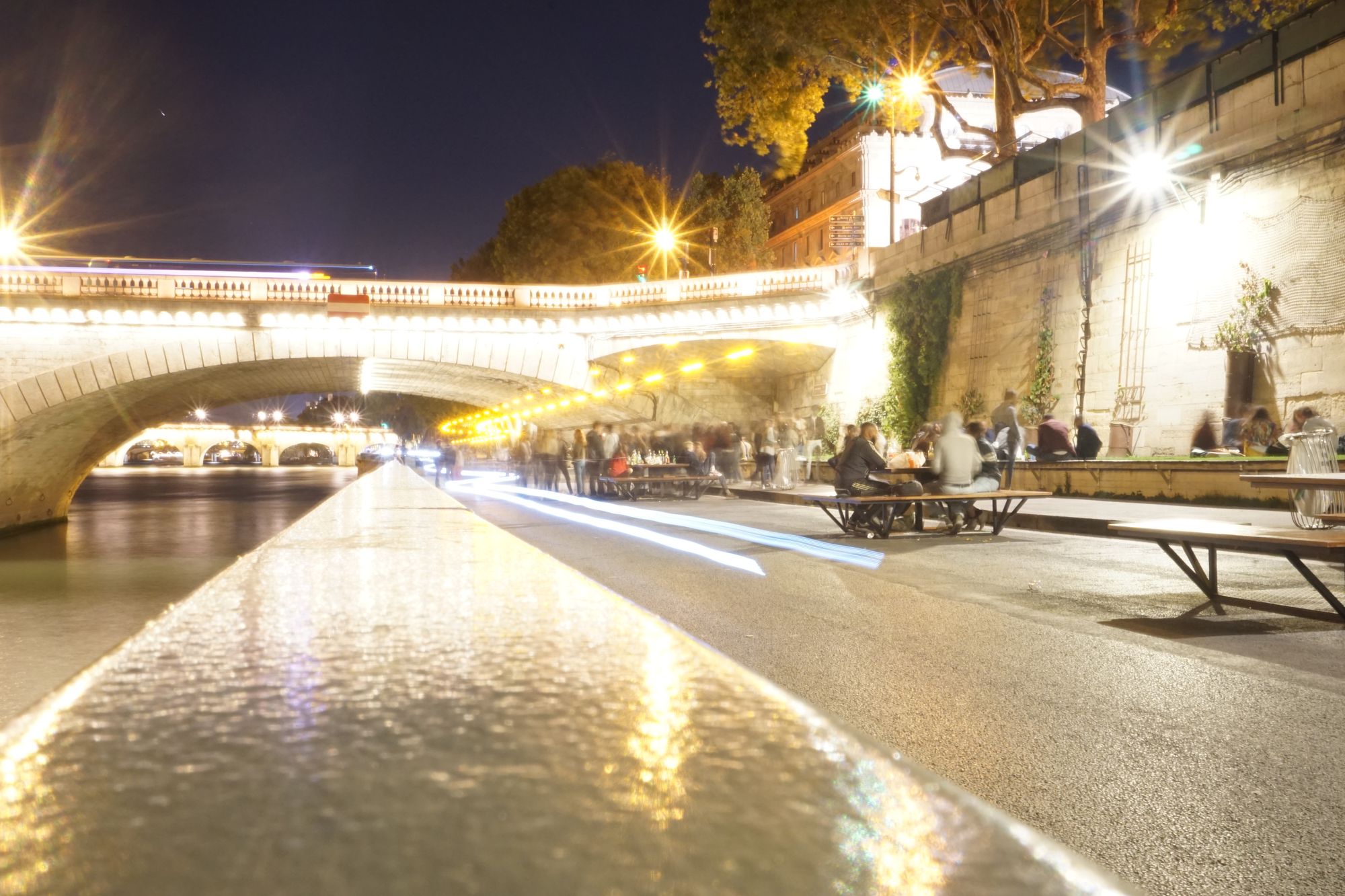 A pedestrianised street by a river at night. People walk and cycle, and warning lights flash on a cleaning truck.