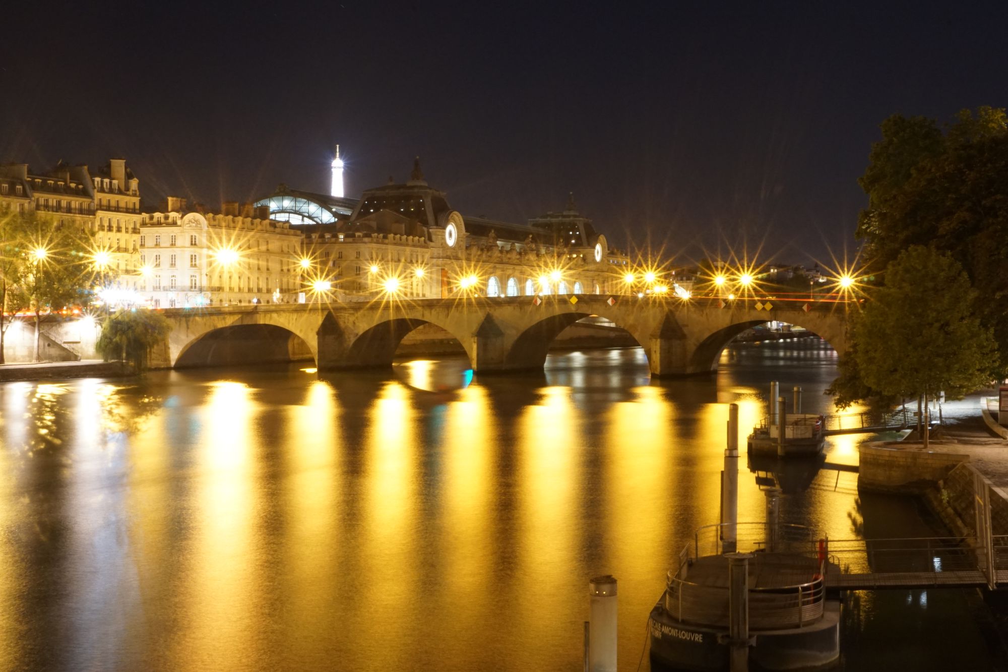 A bridge reflected in a river at night. In the distance, the Eiffel Tower is brightly lit.