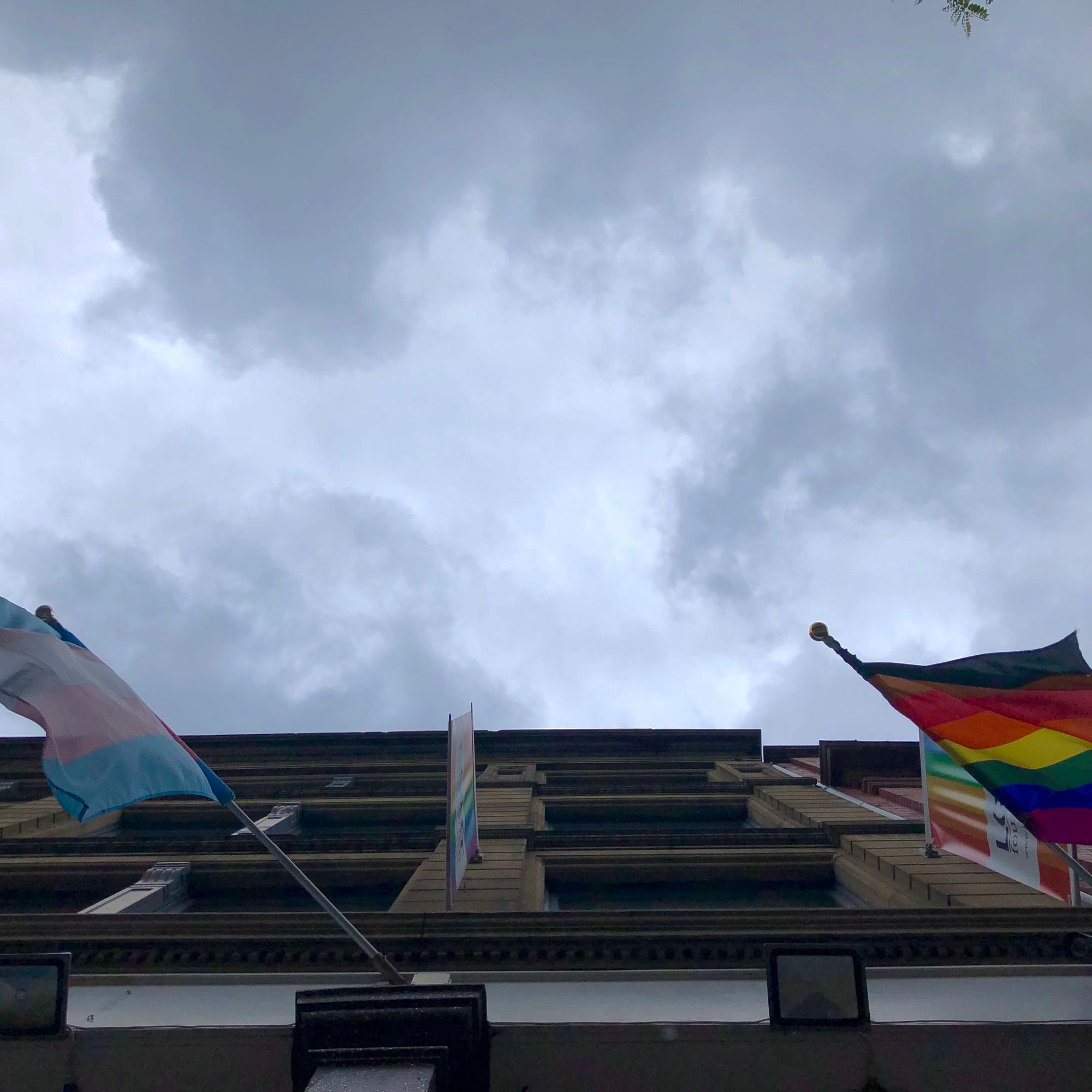 Building frontage with trans pride and gay pride flag (with black and brown stripes)