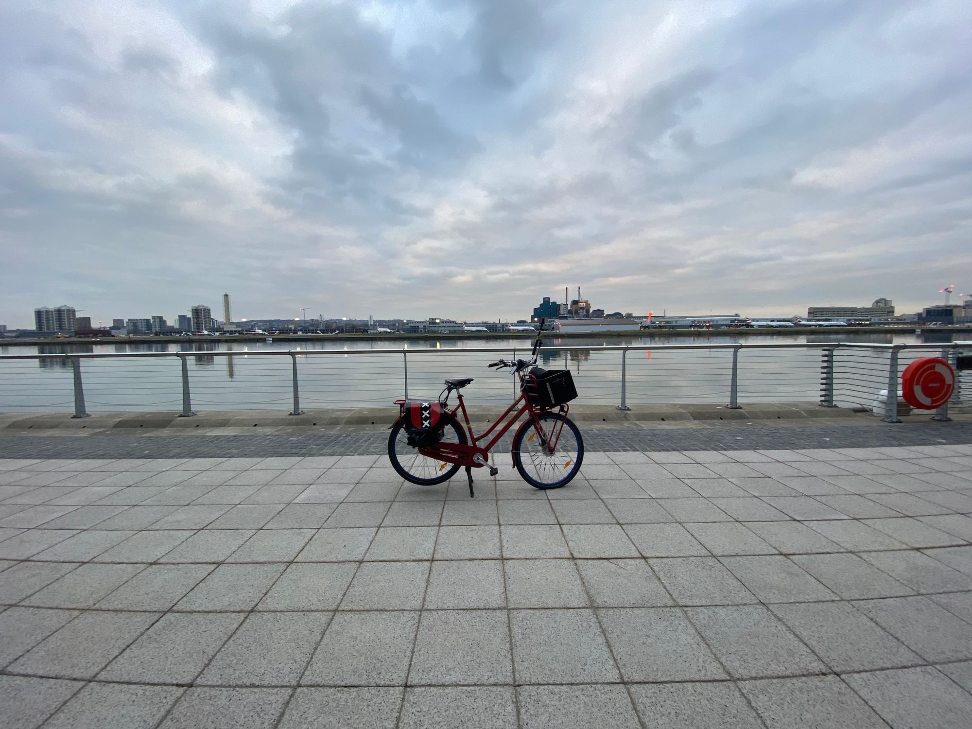 Red Dutch bicycle on a paved area in front of a dock. London City Airport is on the other side.