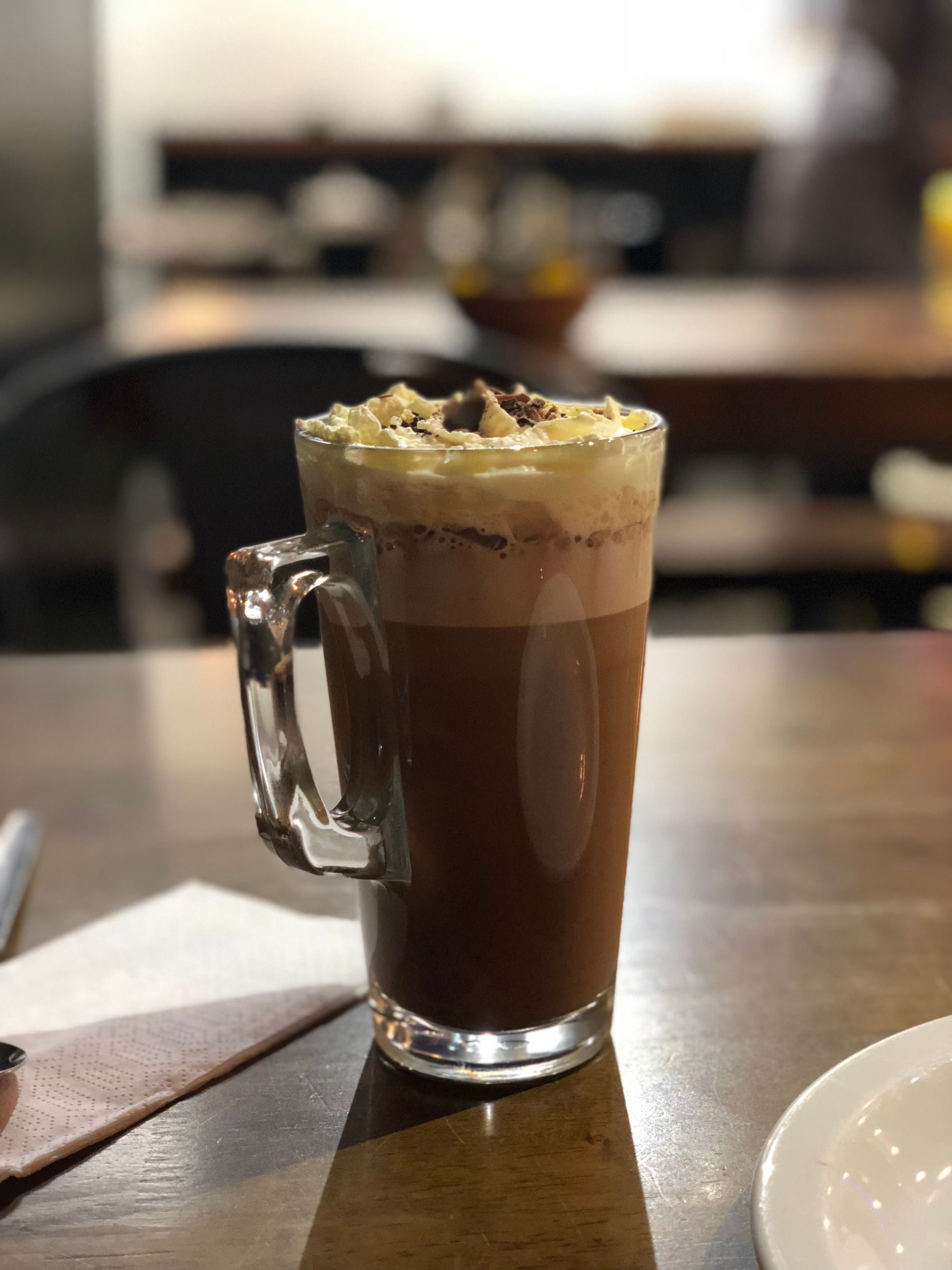 A full glass of coffee with a mocha-like drink, with cream and chocolate shavings on the top, in a coffee shop lit solely with artificial lighting.