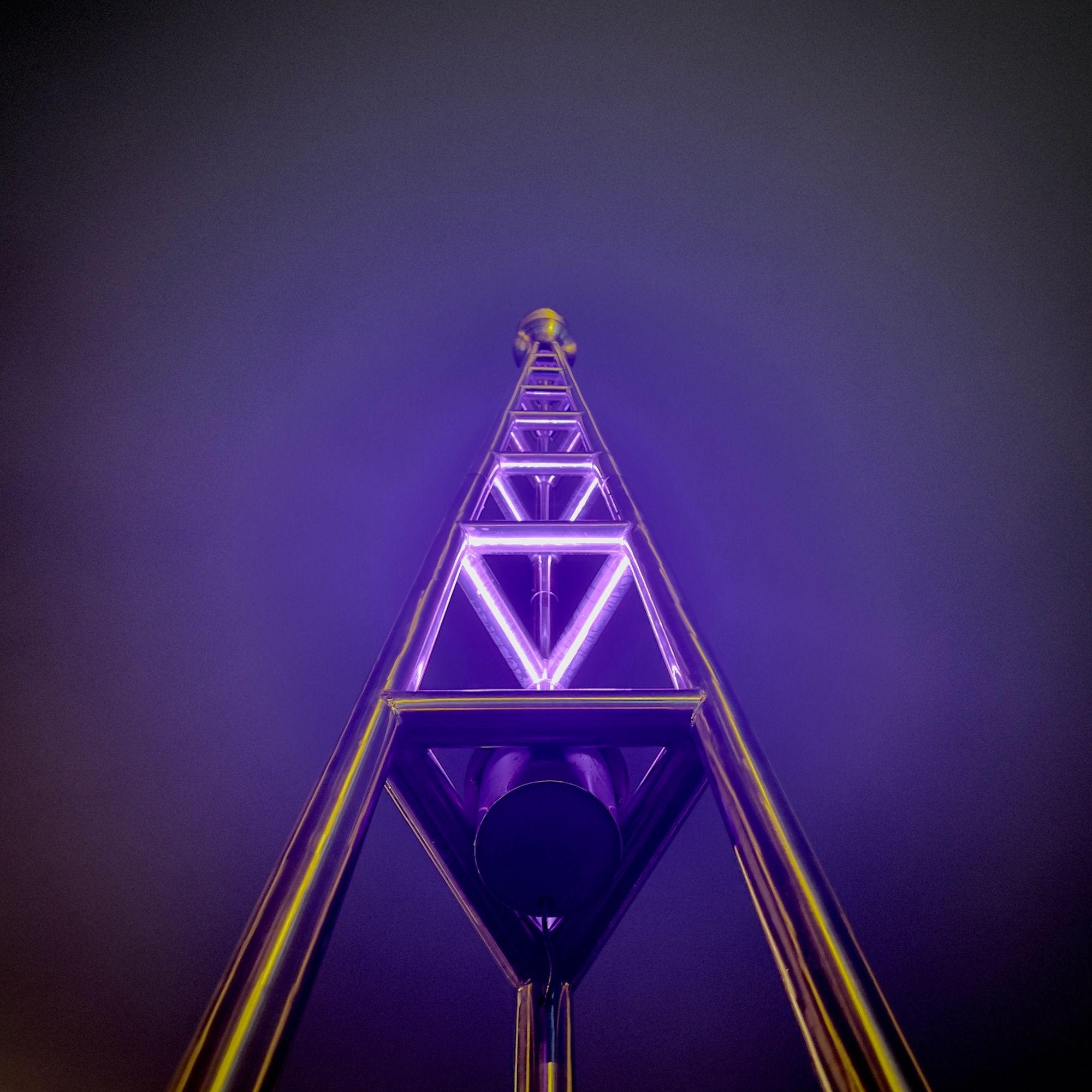 Looking up at a triangular transmitter tower made of shiny metal tubing, with bluish purple tube lights in a triangle arrangement on the tiers.
