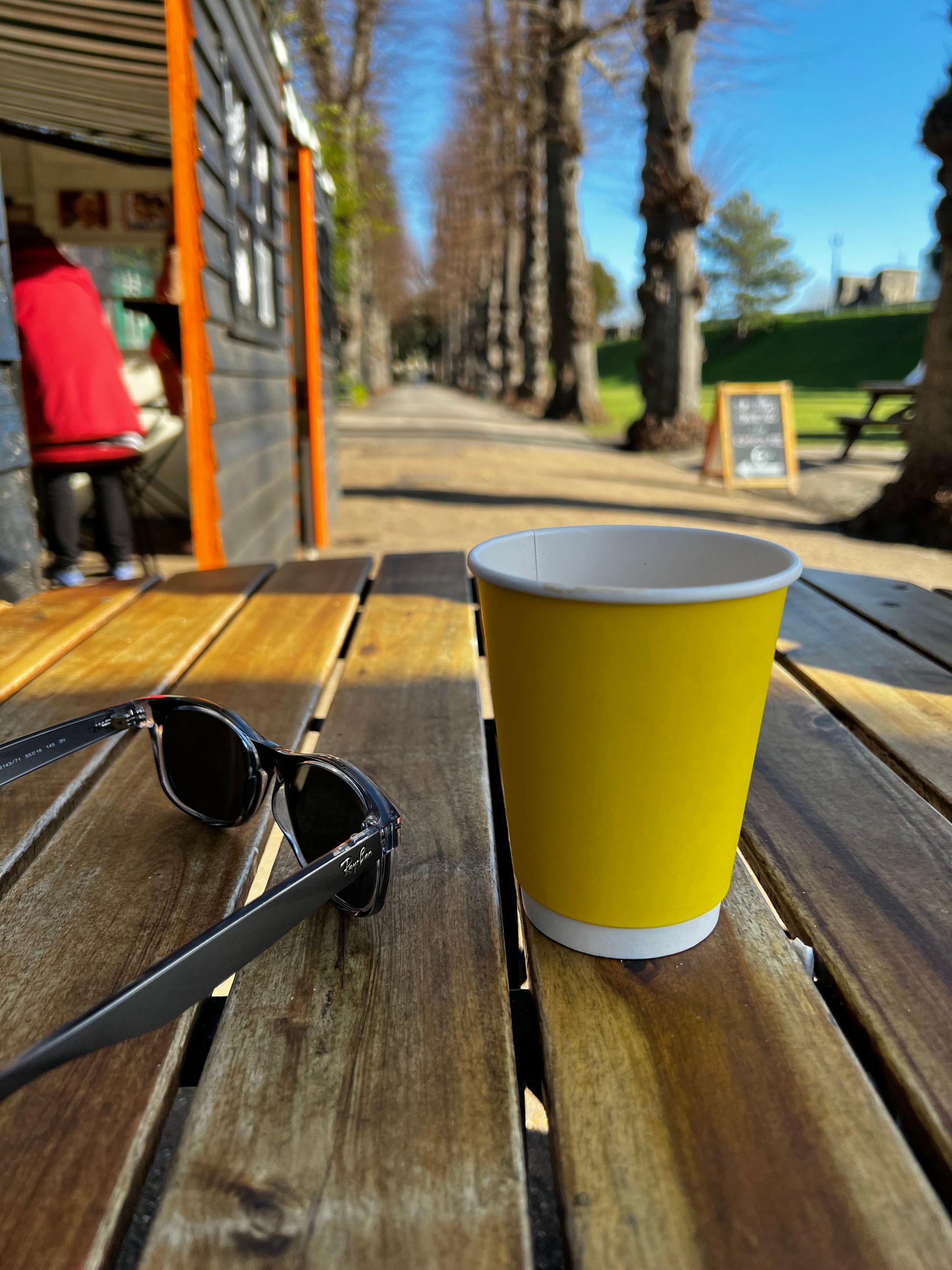 A pair of sunglasses and a yellow takeaway coffee cup on a park table, with a tree-lined avenue and a park café hut.