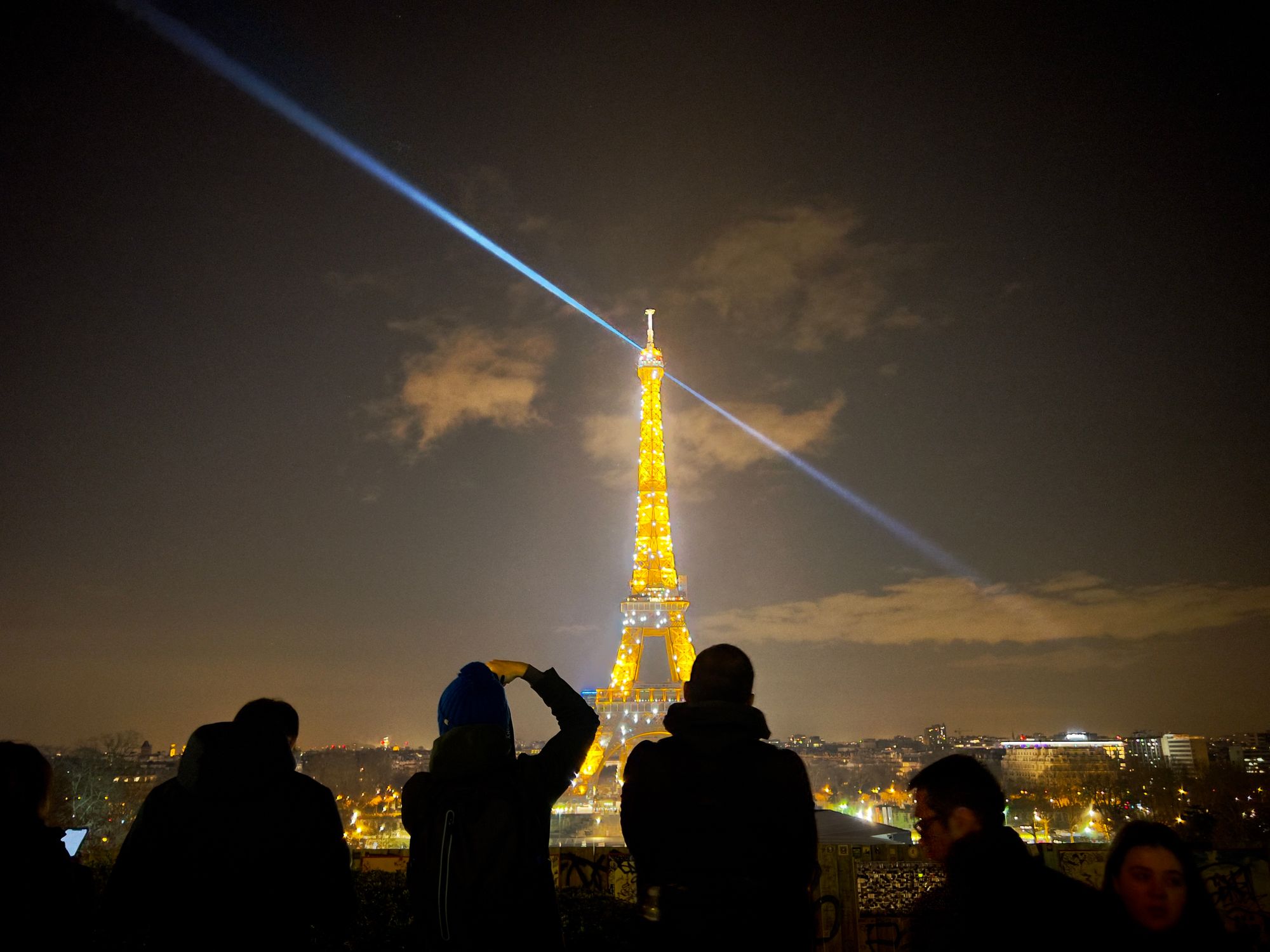 The Eiffel Tower lit up with sparkles at night with its searchlight swinging through the sky. People take photos in the foreground.