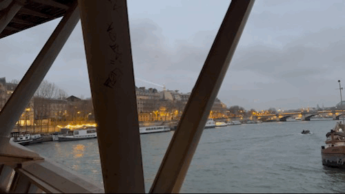 The Eiffel Tower's searchlight sweeps over the Seine at dusk, seen through girders of passerelle Léopold-Sédar-Senghor 