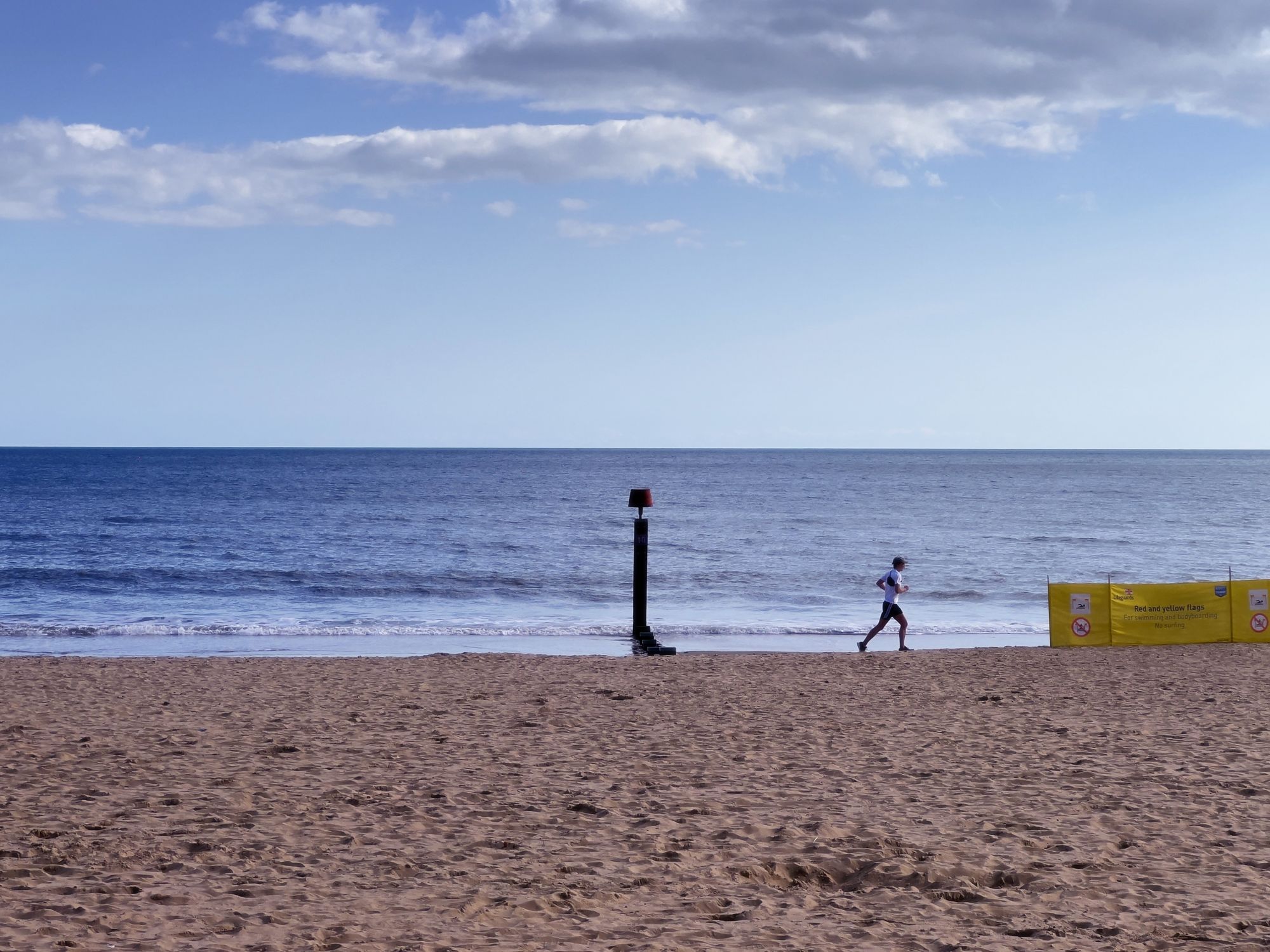 A runner runs past a beach marker on a sunny day, looking out to sea. A yellow fabric barrier carries a warning about red and yellow flags.