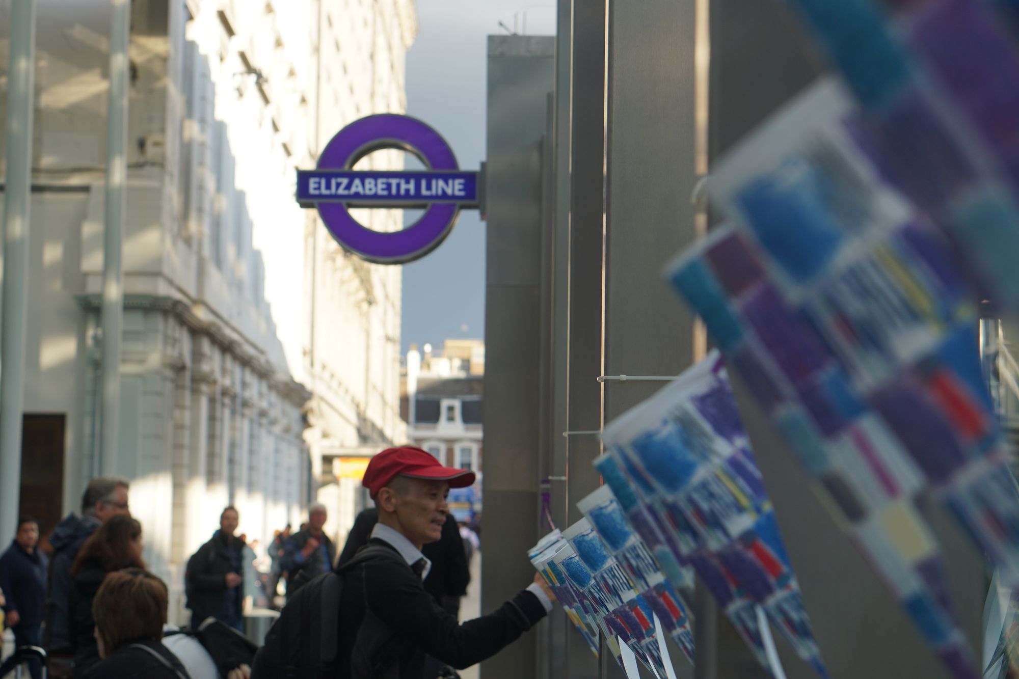 Bunting with blue and purple kids' drawings on it is strung up from pillars outside an Elizabeth Line station, with a man in a red cap examining them closely.