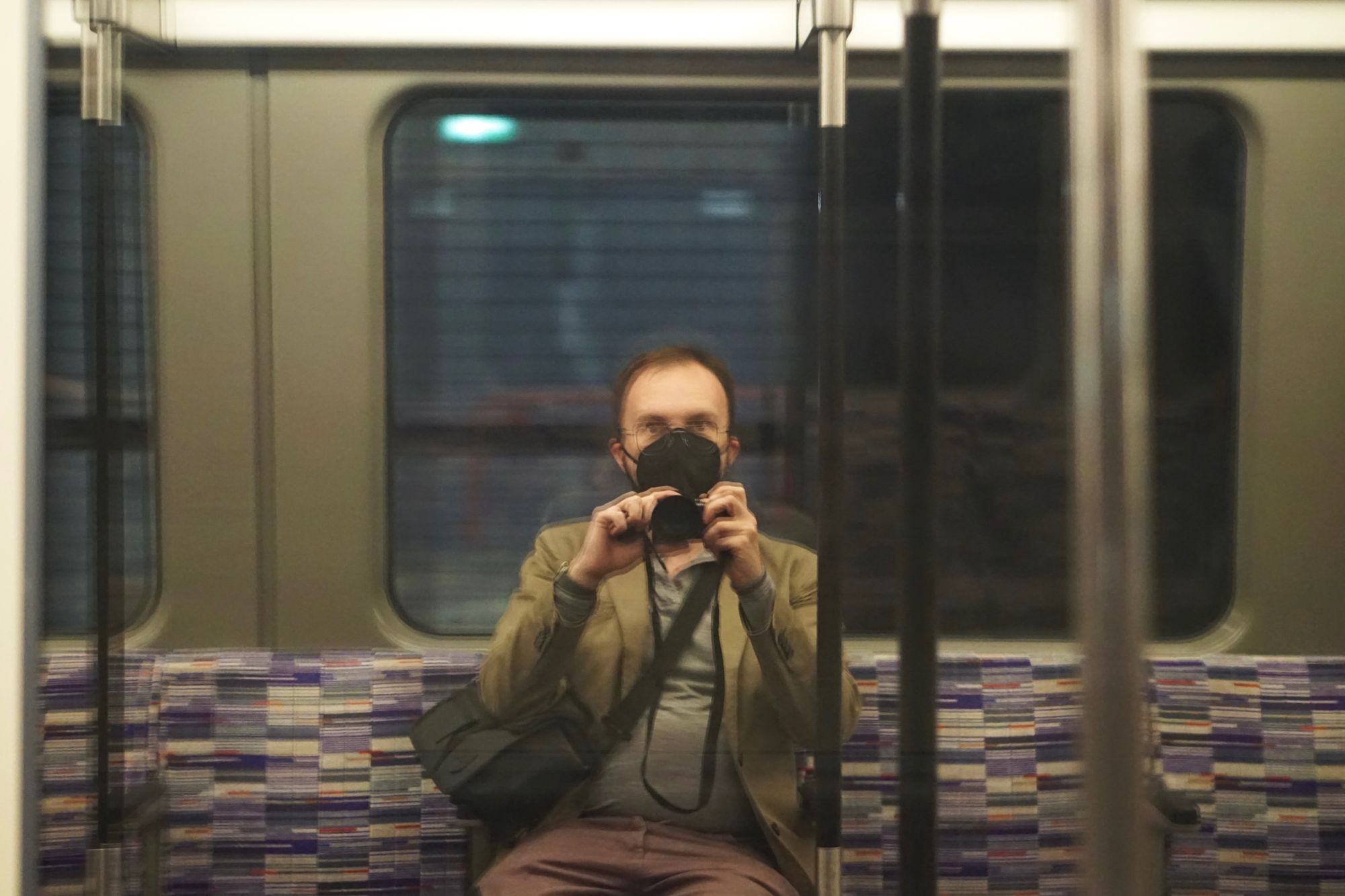Jonathan taking a photo of his own reflection in the train window, wearing a mask and a cream coloured blazer.