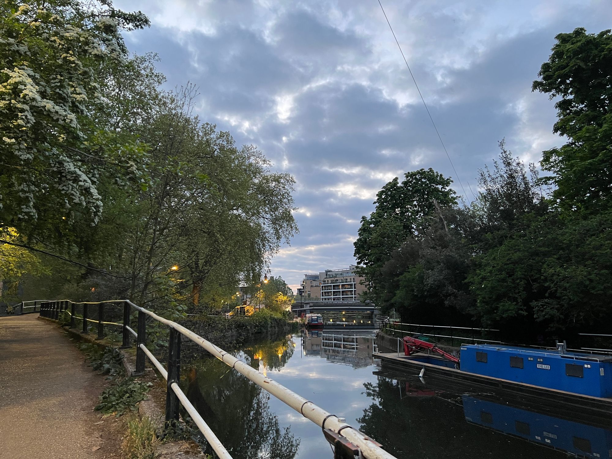 A canal at dusk with lights on the towpath, trees in white blossom, and a blue boat moored.