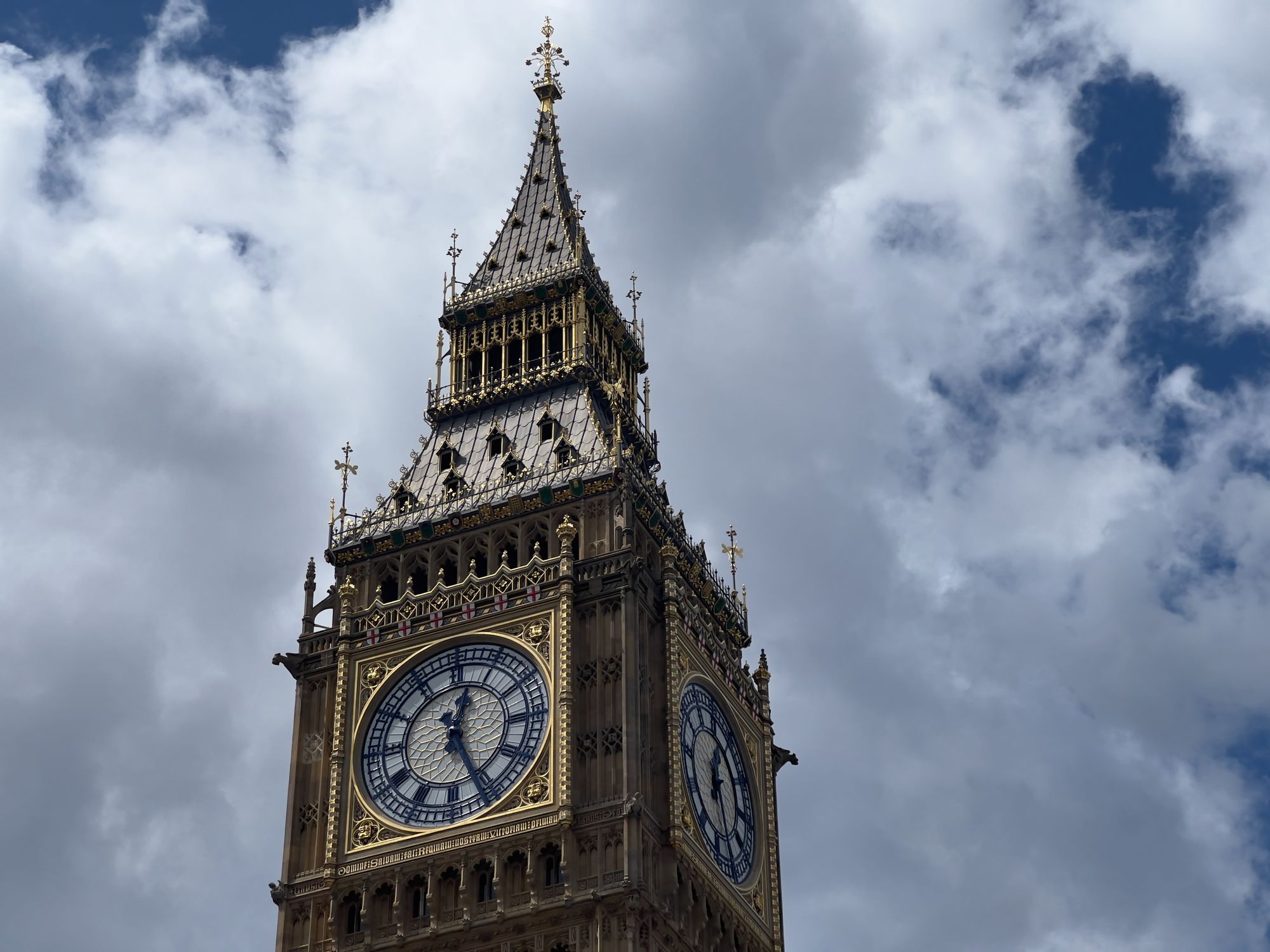 The top of the Palace of Westminster clock tower (Big Ben), in gilded gold and blue, against a partly cloudy sky.