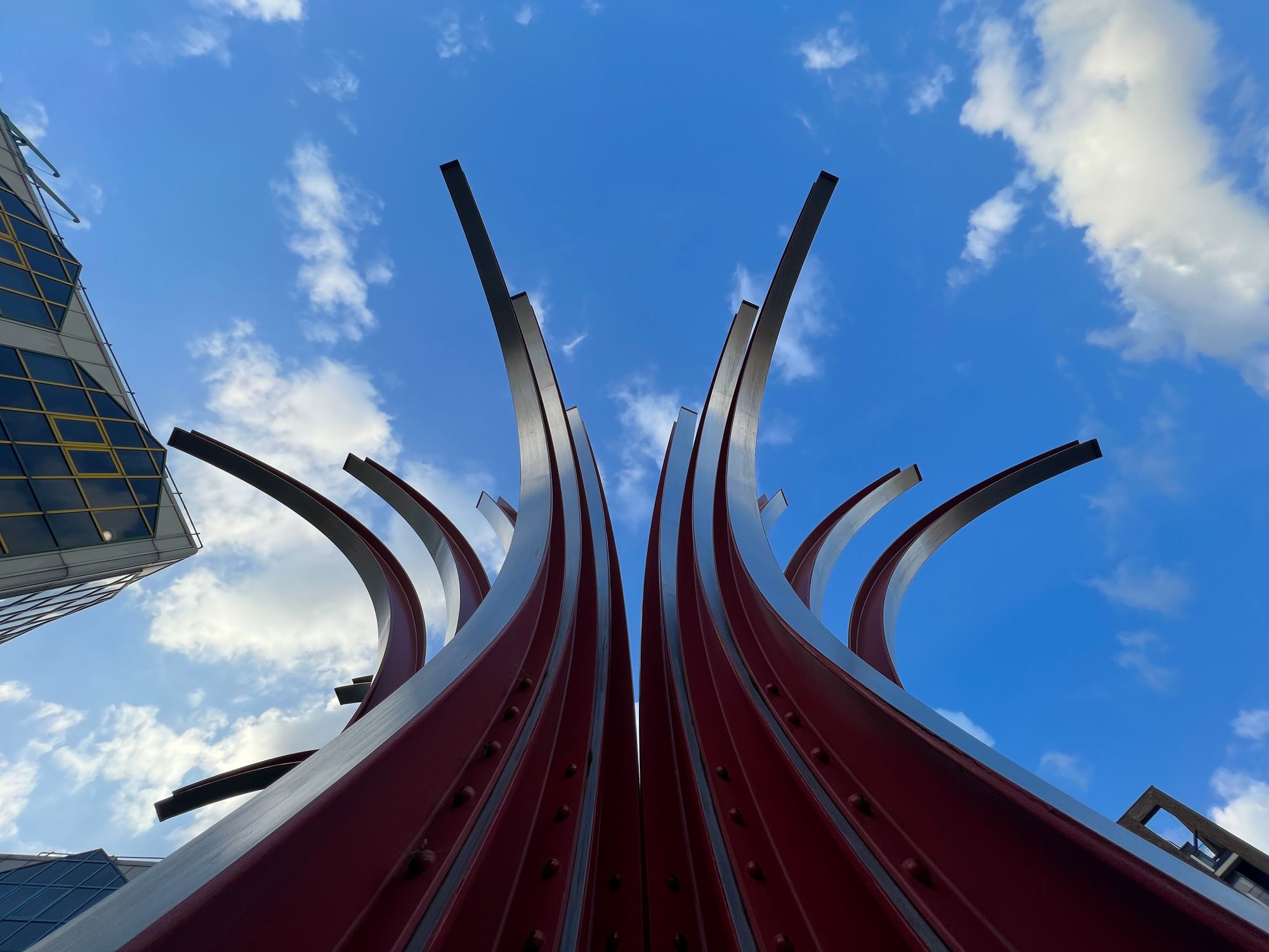 A sculpture made of rails curving upwards in a bunch rises into the sky amongst tall buildings.