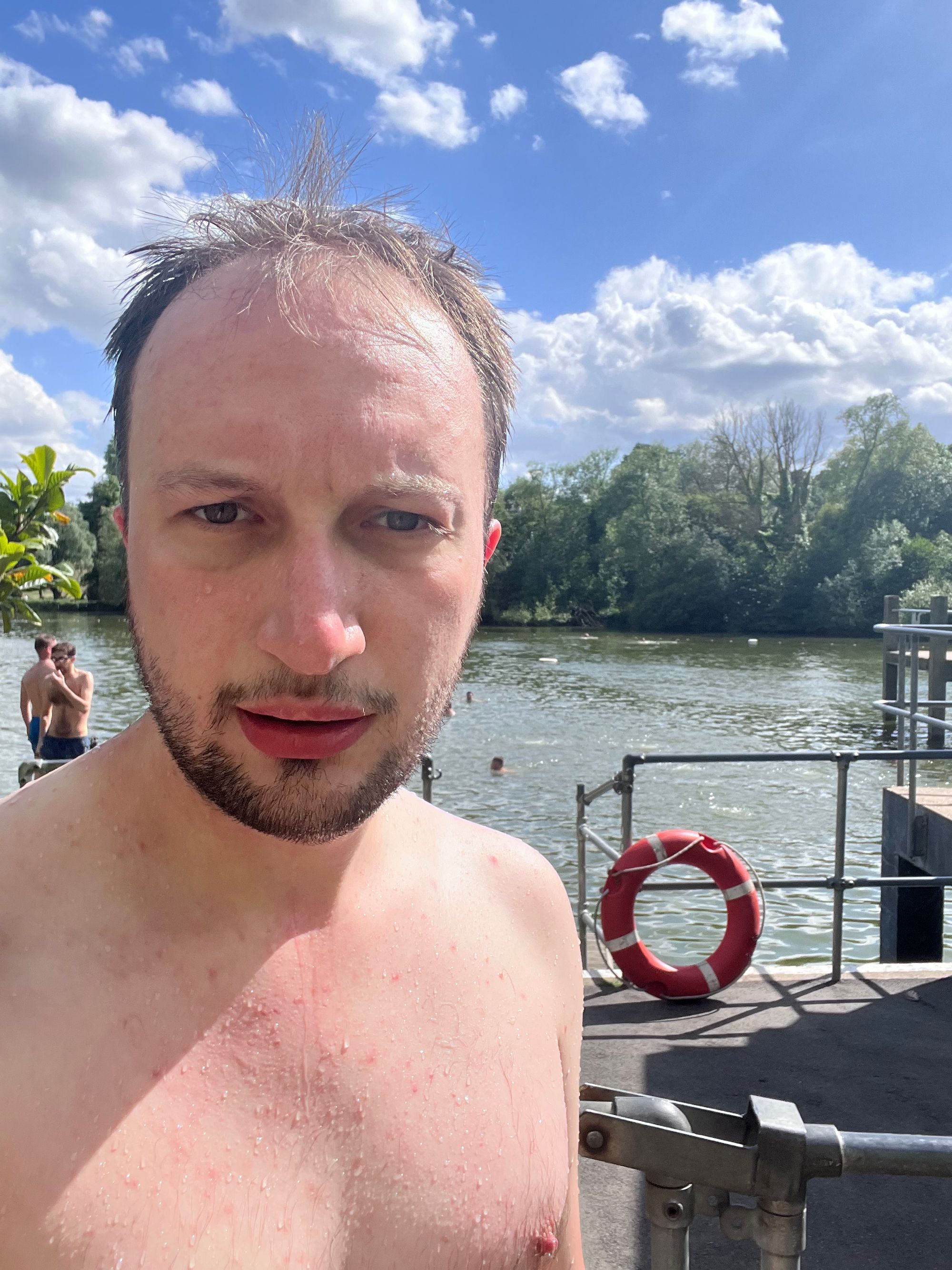 Jonathan stand sin front of a pond with people swimming in it. He wears no shirt and is covered in water droplets.