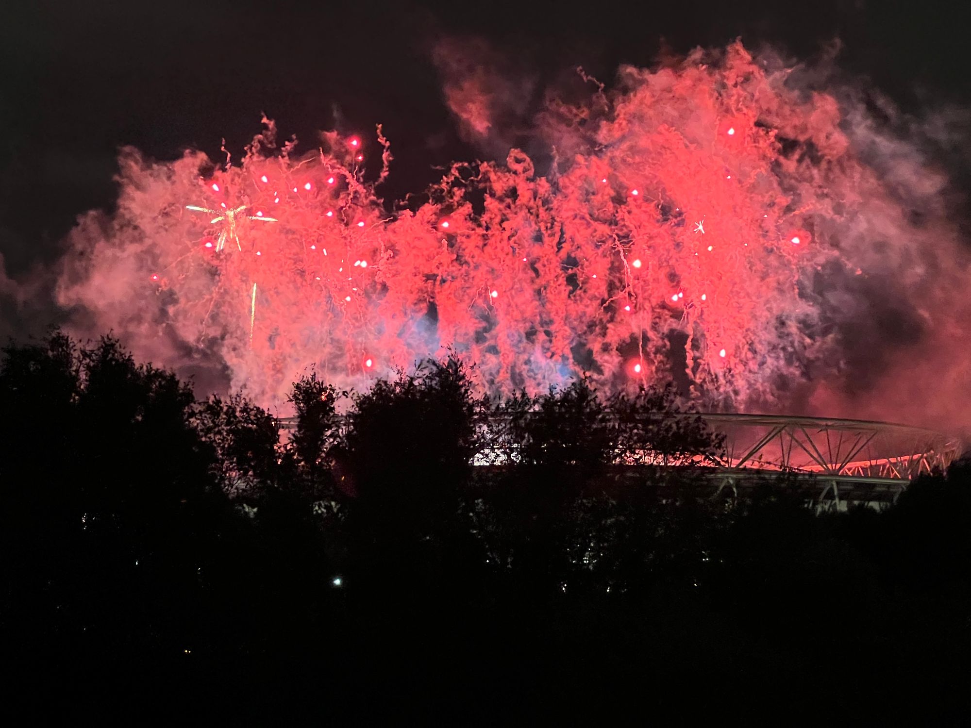 The London Stadium at night in silhouette, with red pyrotechnics above it lighting smoke from previous explosions.