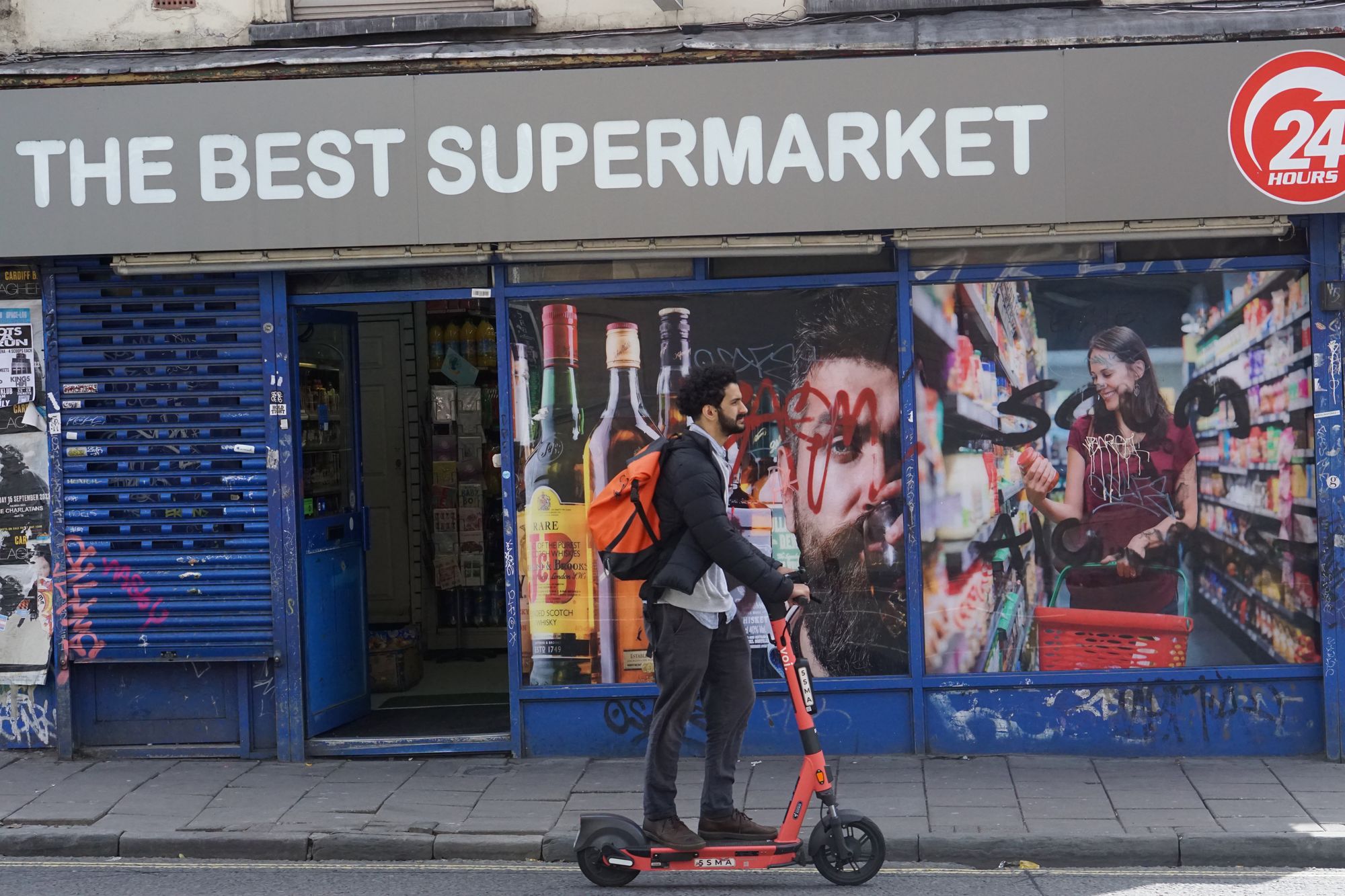 A man with a beard and an orange backpack rides an electric scooter in front of a graffitied shopfront for "The Best Supermarket."
