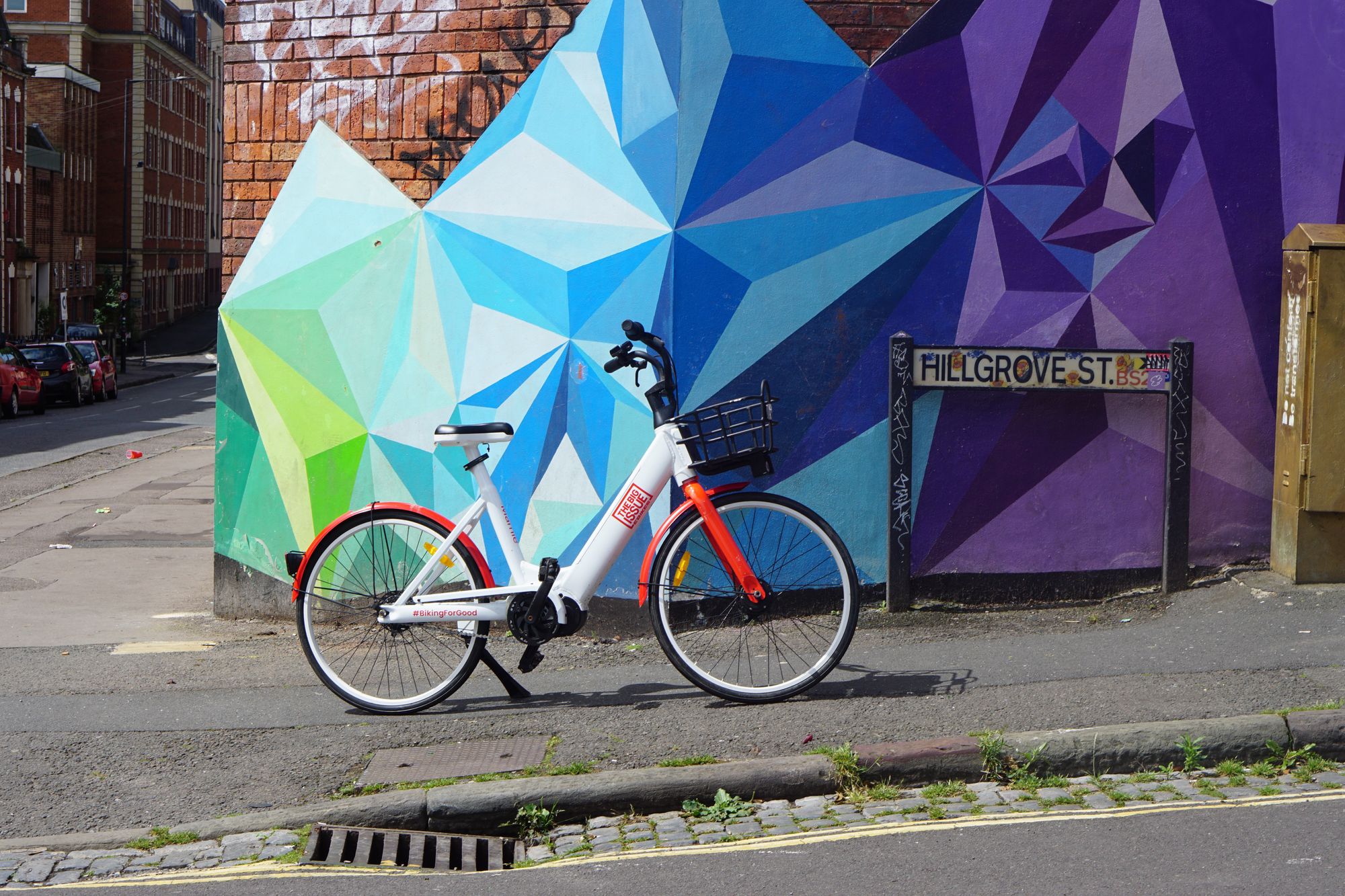 A Big Issue-branded rental bike propped on a stand in front of a wall decorated with a colourful geometric pattern on Hillgrove Street.