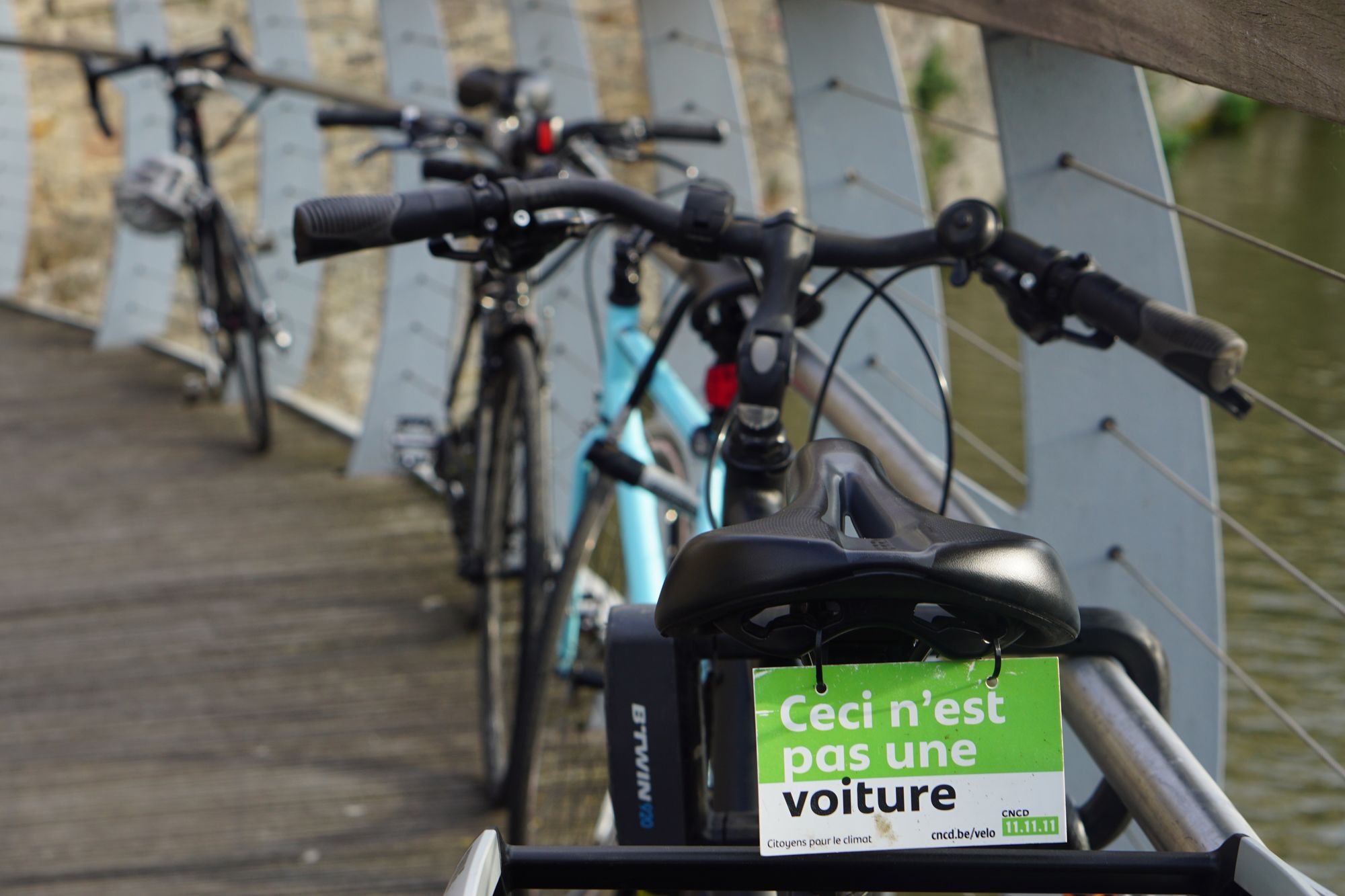 The rear saddle of a bicycle parked on a bridge with railings. Hanging from the saddle: "ceci n'est pas one voiture."