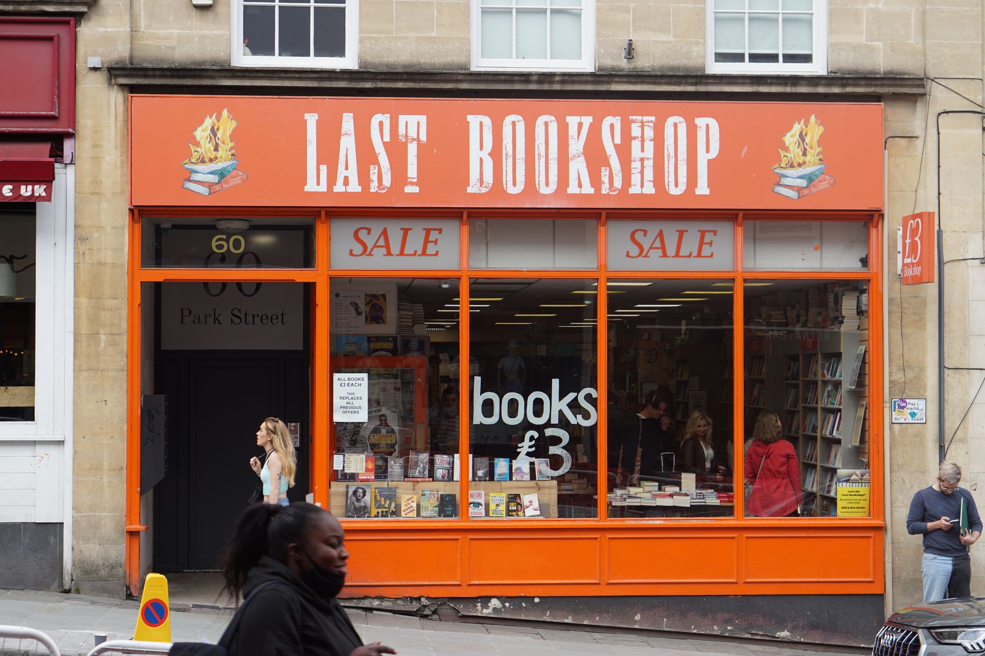 People walk past the orange shopfront for "Last Bookshop", with a logo of burning books and the words "SALE. Books £3"