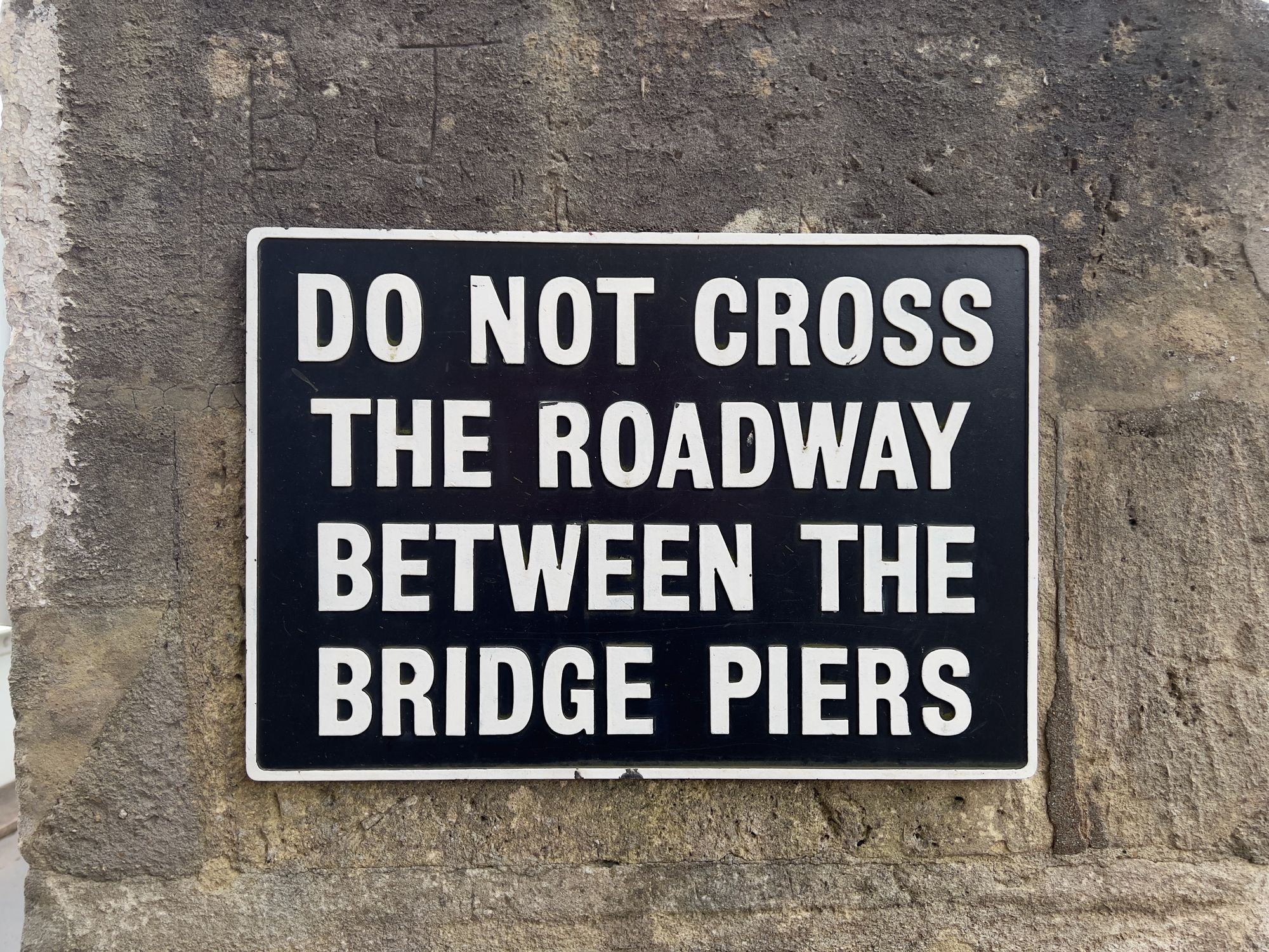 A white-on-black sign on a stone wall: "DO NOT CROSS THE ROADWAY BETWEEN THE BRIDGE PIERS." The letters BJ are carved above.