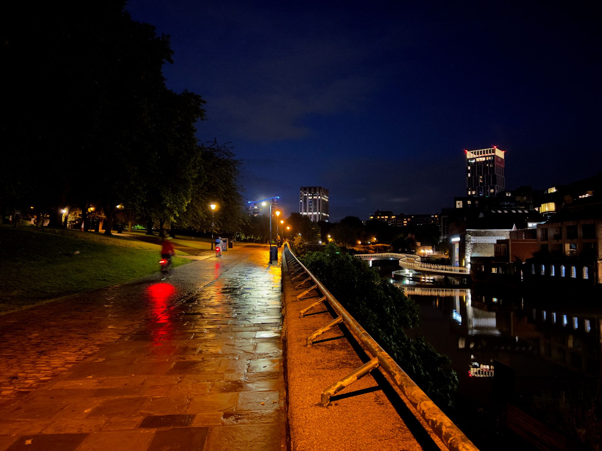 A riverside footpath and cycle path with cyclists riding away, with a park to the left and a twisty bridge over the river on the right.