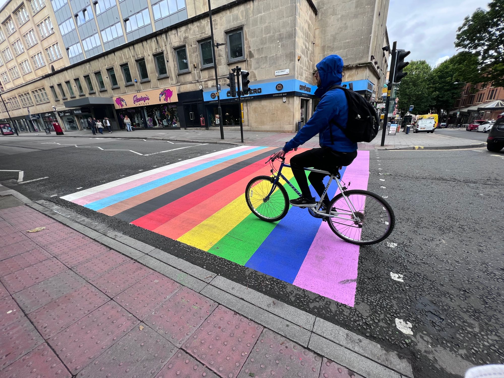 A man cycles past over a pedestrian crossing (lighted), marked out with the stripes of a trans- and race-inclusive pride flag.