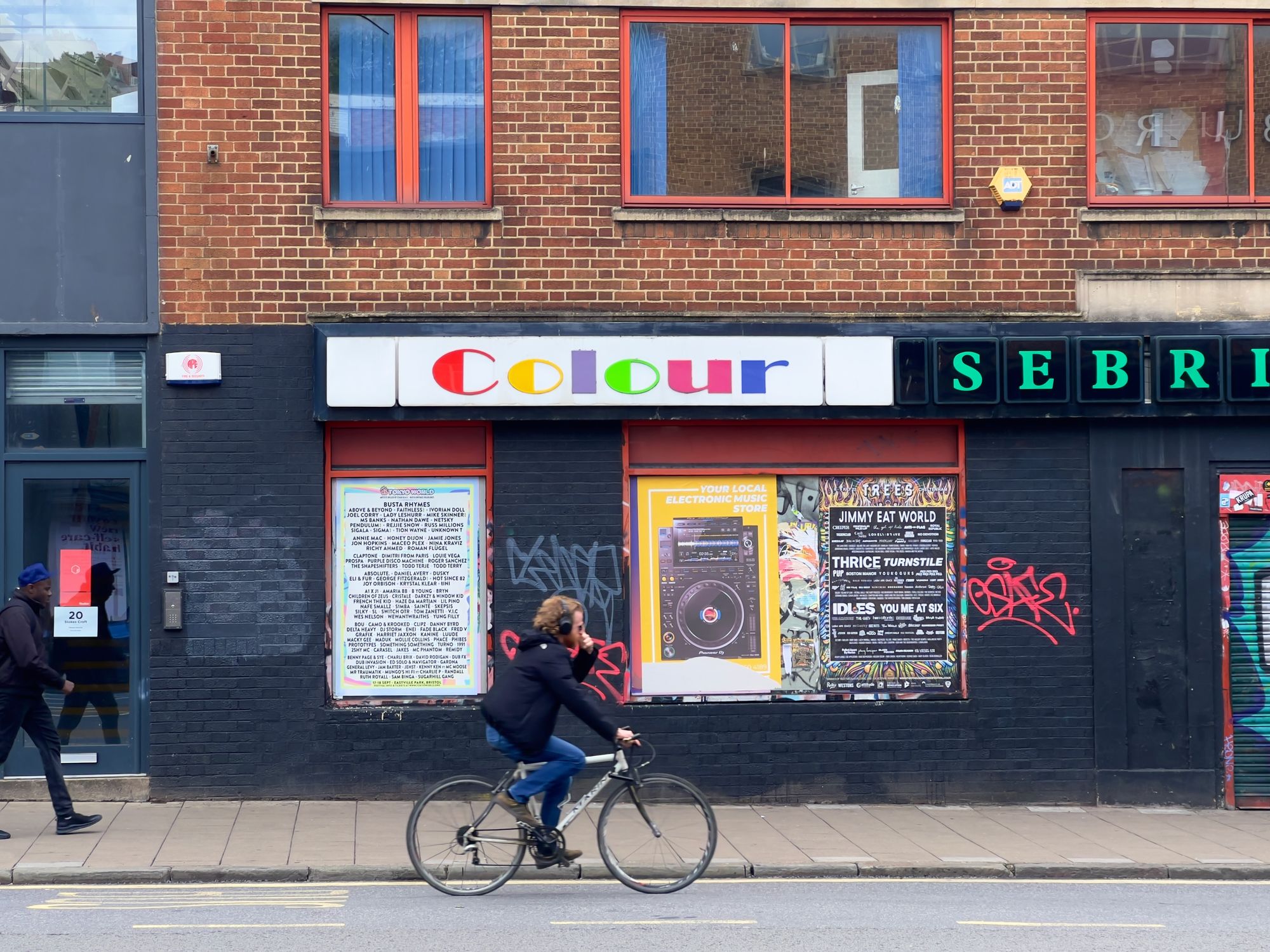 A man cycling wearing headphones in front of a colour printers' shop with bills posted on the side.