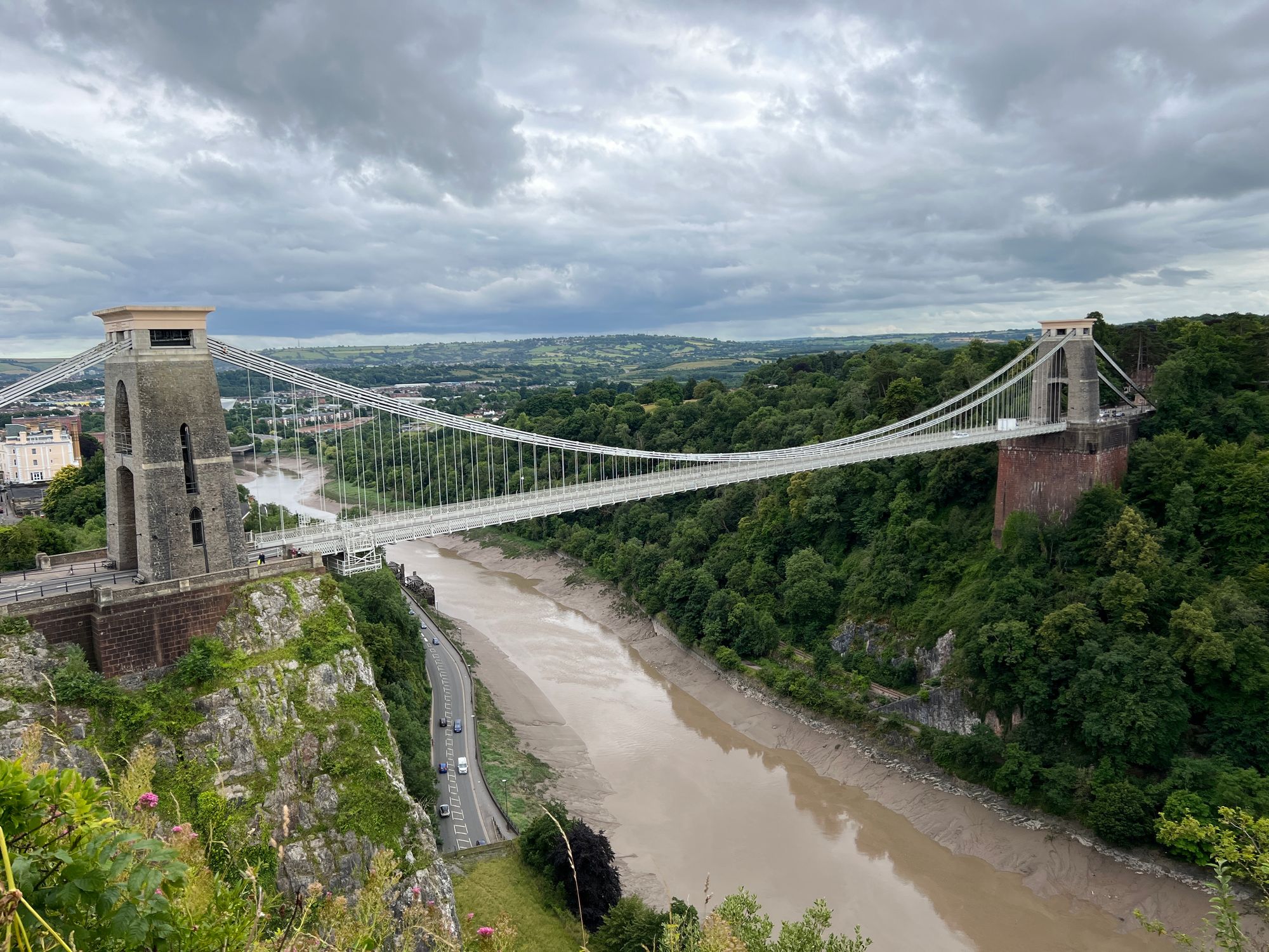 A suspension bridge with two brick towers across a wide gorge with the river tide coming in.