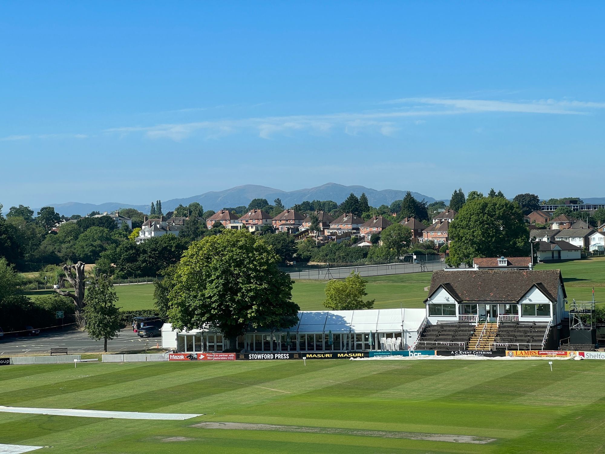 A view out over a cricket field with houses and fields behind it, and some imposing hills in the distance.