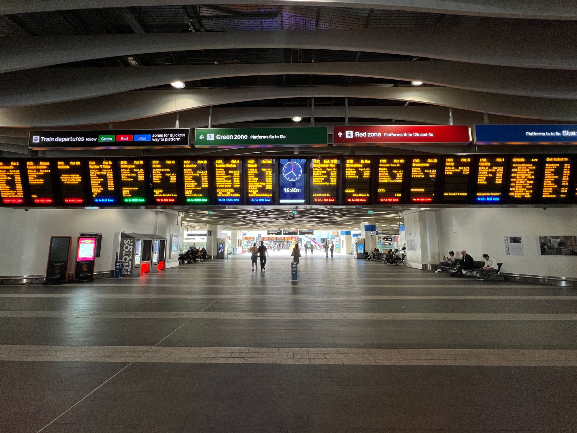 A passageway through to a concourse at a railway station with a tiled floor, a row of departures boards, and signs to the Blue, Red, and Green Zone.