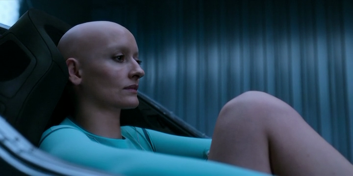 The Halsey clone, a 40-something woman with a bald head wearing a tunic, sits in a horizontal sleeping pod.