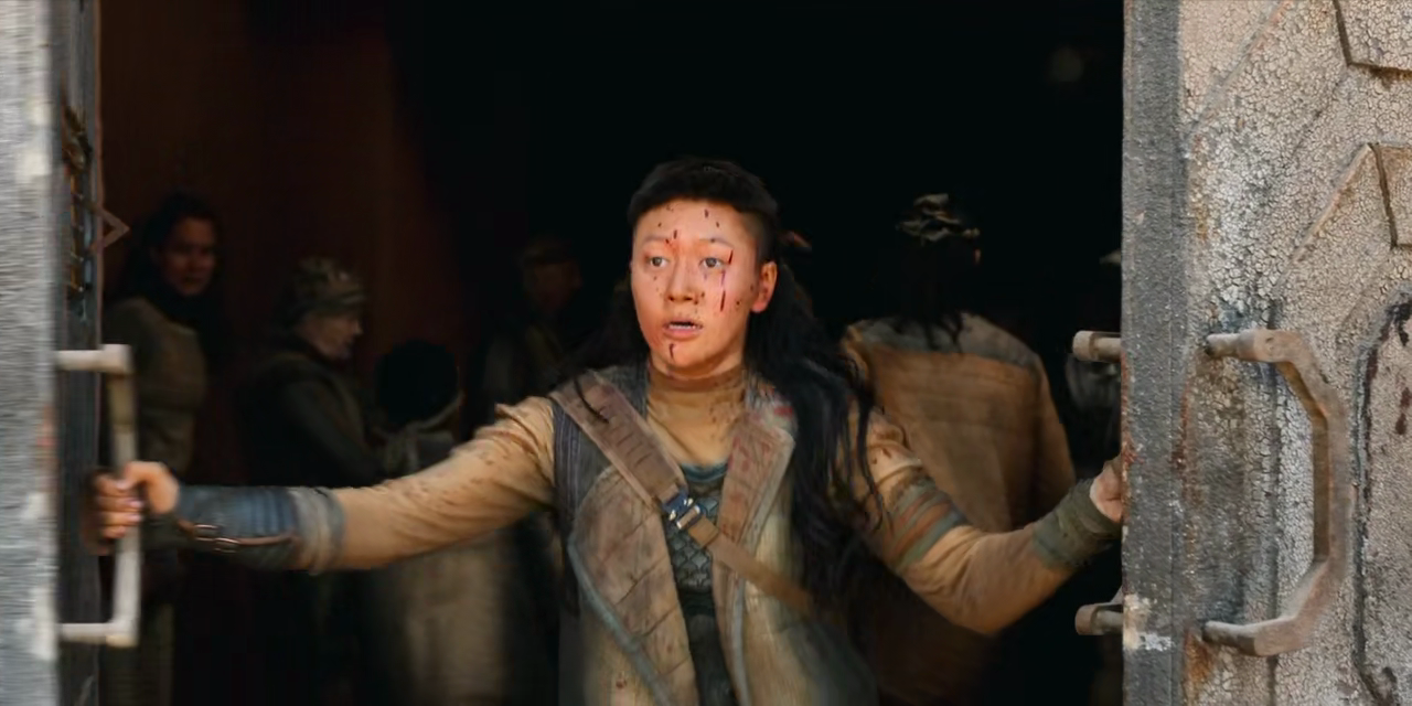 Kwan Ha, a young woman with blood spatters on her face wearing utilitarian brown clothing, shuts some bulkheads with people inside.