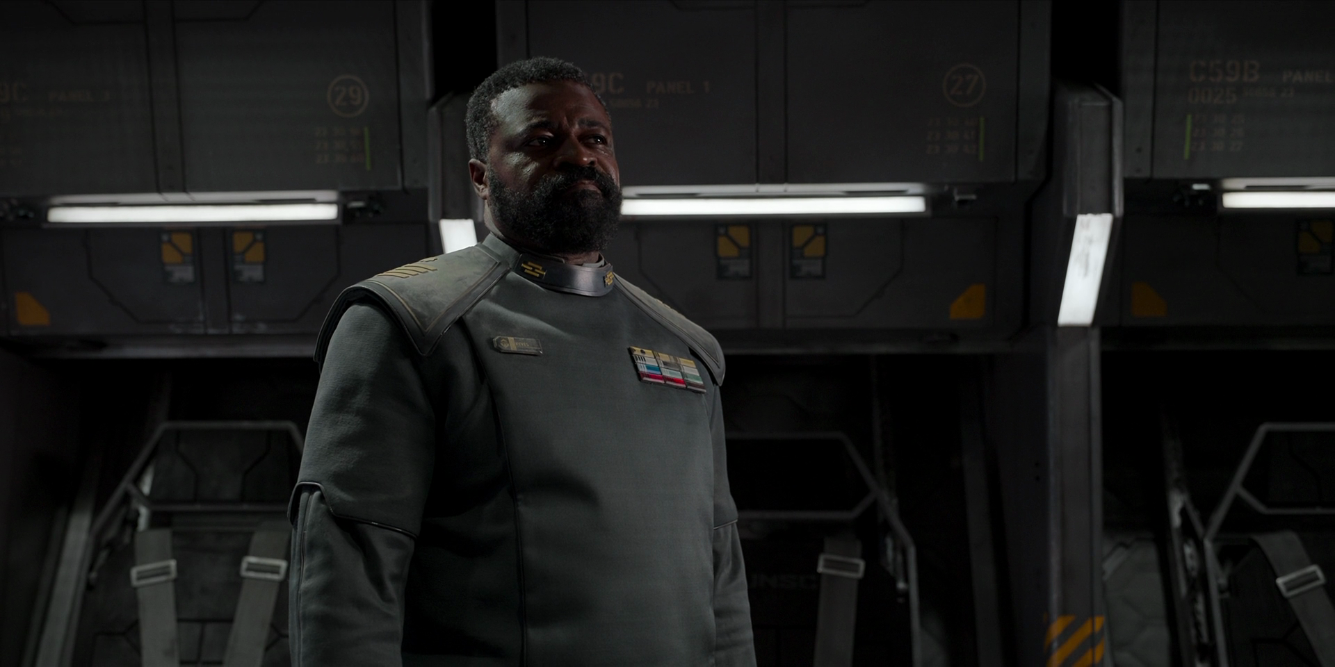 Captain Keyes, a Black man with a beard wearing a grey navy tunic, stands in a military spacecraft's passenger bay.