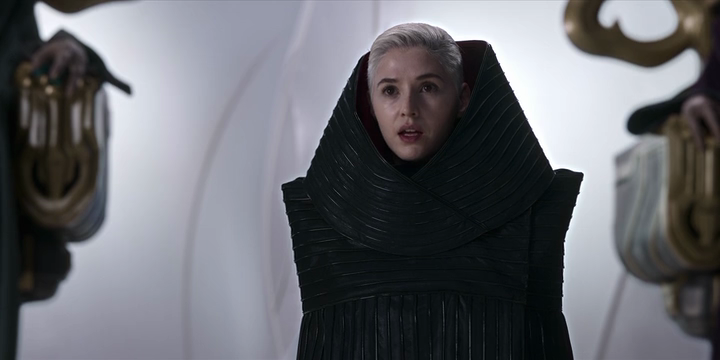 Makee, a white woman with short platinum-blonde hair, wears a rectangular black oversized dress with an enormous collar.