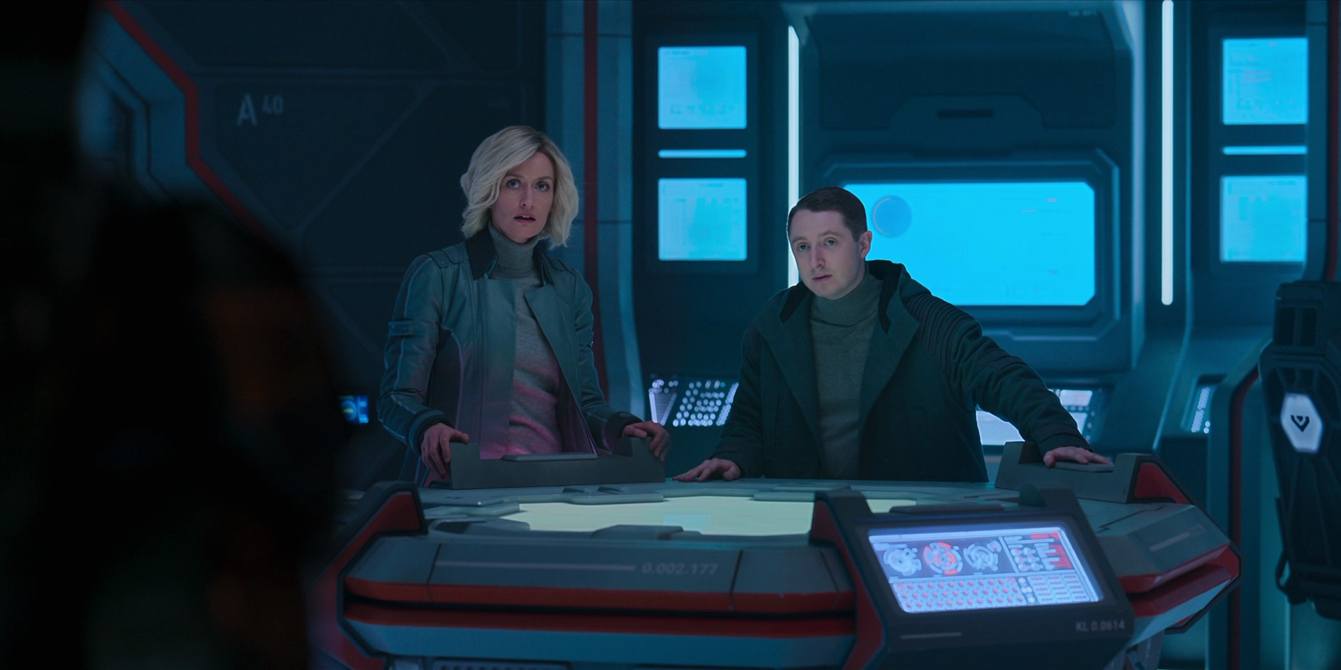 Dr Halsey, a blonde white woman in a grey jacket, and Adun, a shorter white man with dark brown hair, stand behind a console, looking at the Master Chief.