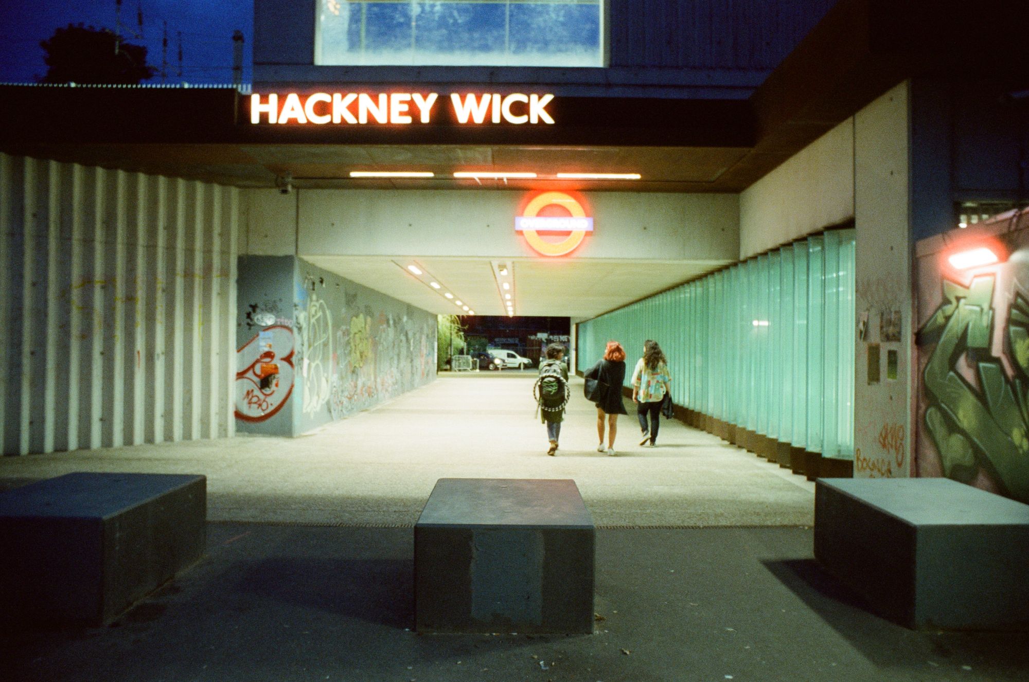 Three people walk away from camera through an underpass to Hackney Wick station. The illuminated lettering and roundel have a fizzy glow like a lightsaber.