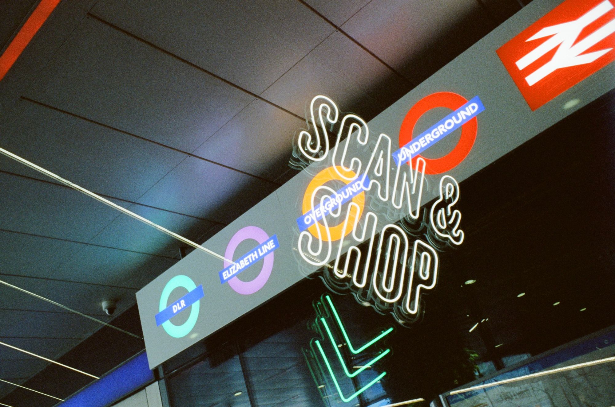 An illuminated station sign with roundels for DLR, Elizabeth Line, Overground, Underground, and British Rail. Exposed over the top: the words "SCAN AND SHOP" in neon lettering.