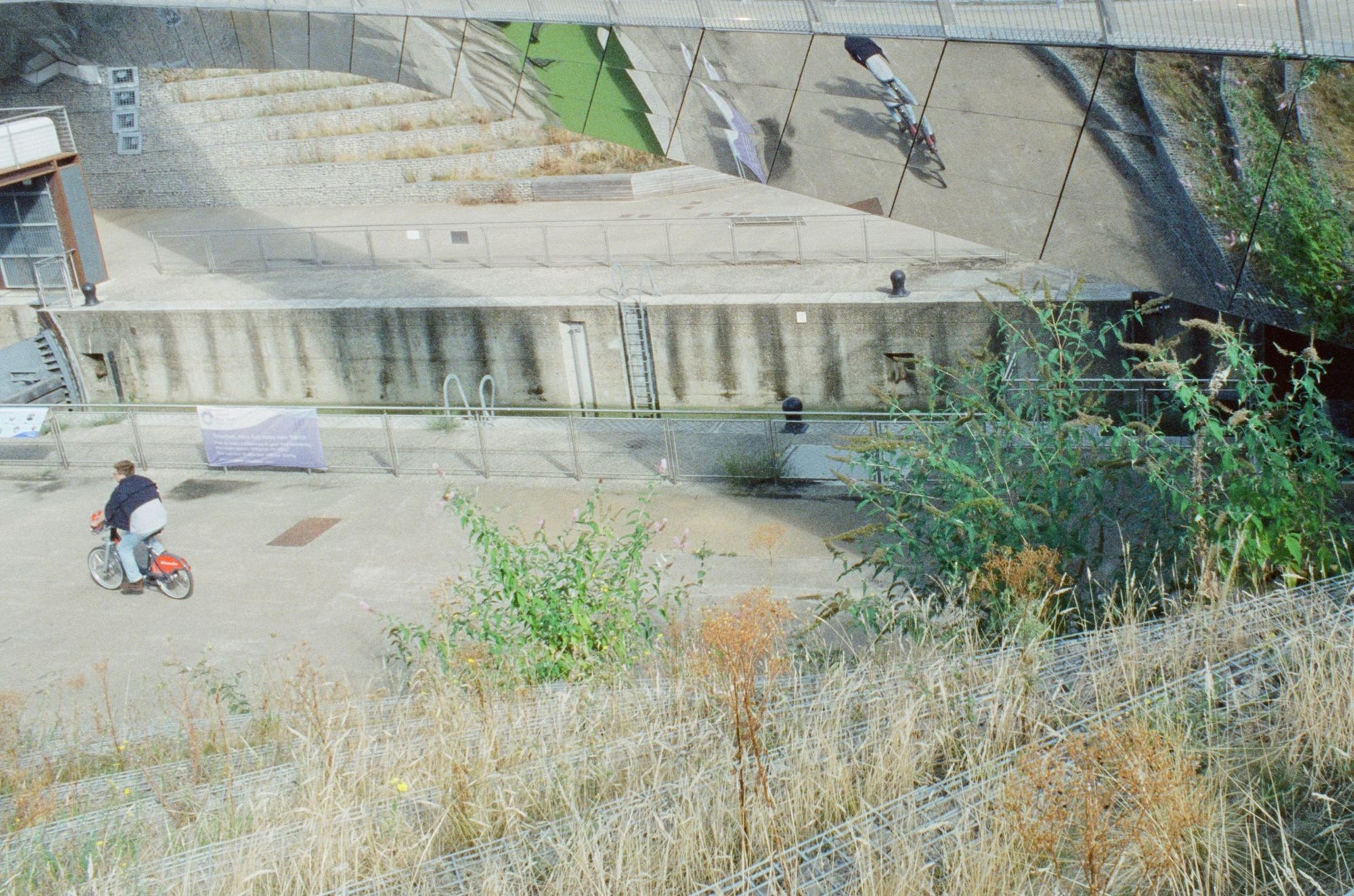 Down onto a canal towpath as someone cycles away on a hire bike. They are reflected in a curved bridge soaring above with reflective panels on the underside.