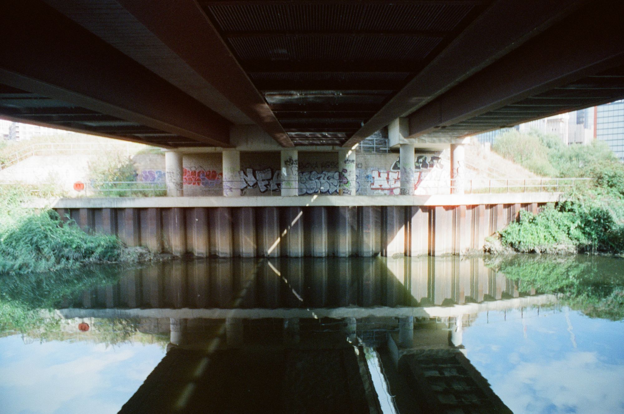 Across a canalised river under a road bridge, with the sky and the bridge reflected in the water. There is graffiti on the far wall.