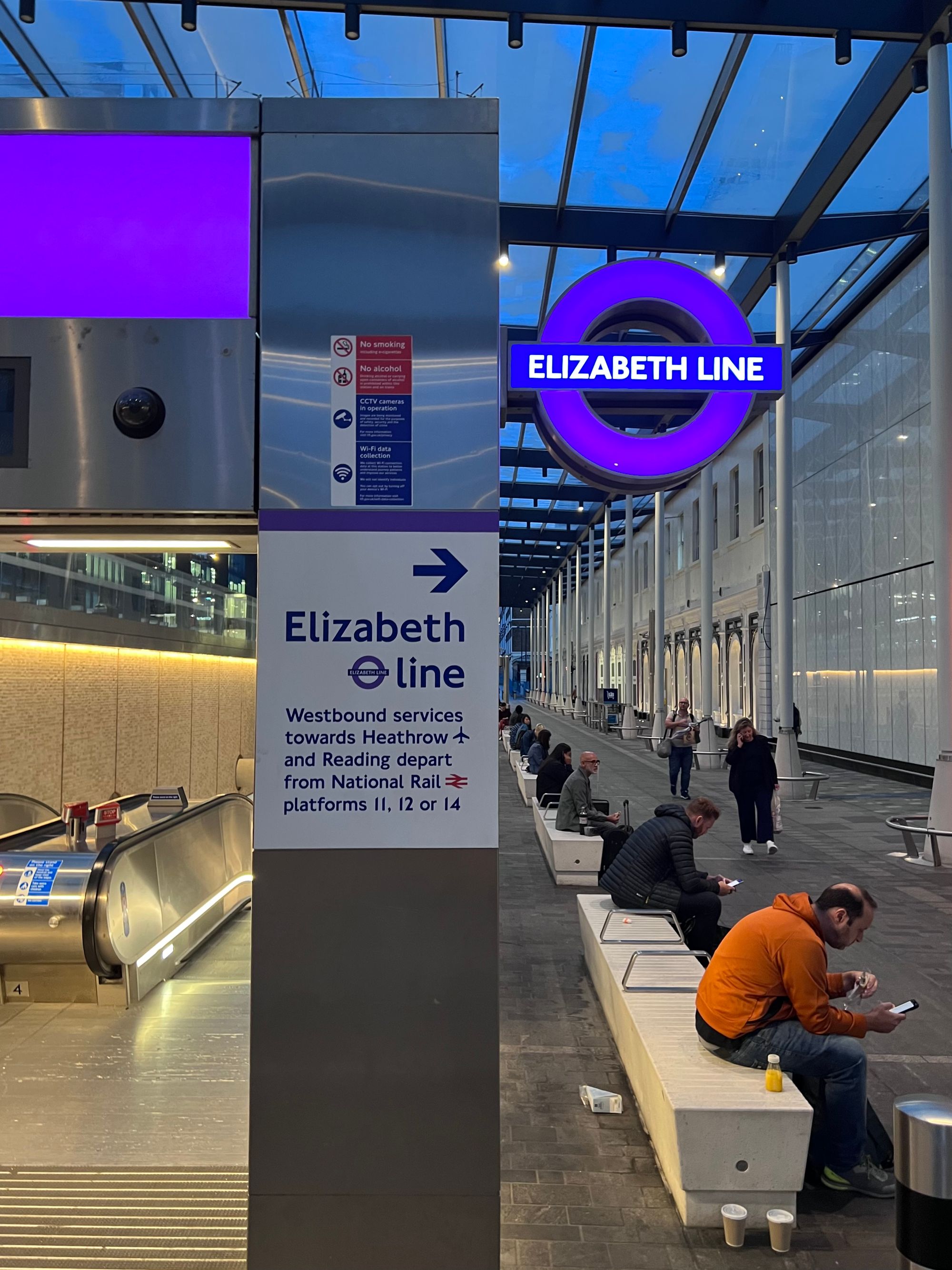 Looking along a covered colonnade from the entrance arch to Paddington Elizabeth line station, its purple roundel glowing.