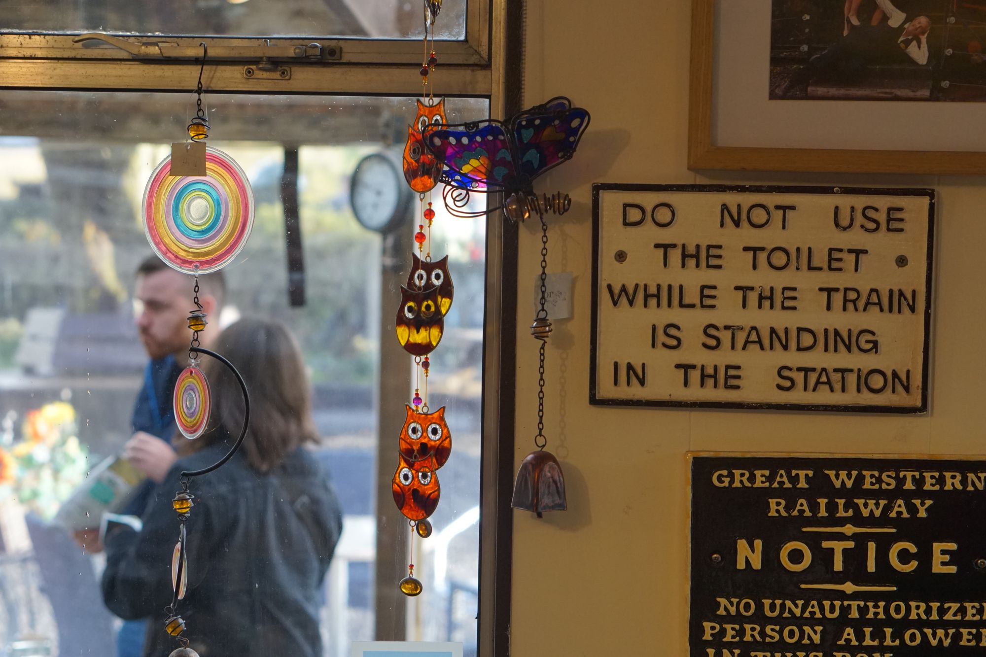 Charms of owls and butterflies hang in the window of a shop, with an old railwayana sign stuck to a wall: "DO NOT USE THE TOILET WHILE THE TRAIN IS STANDING IN THE STATION"