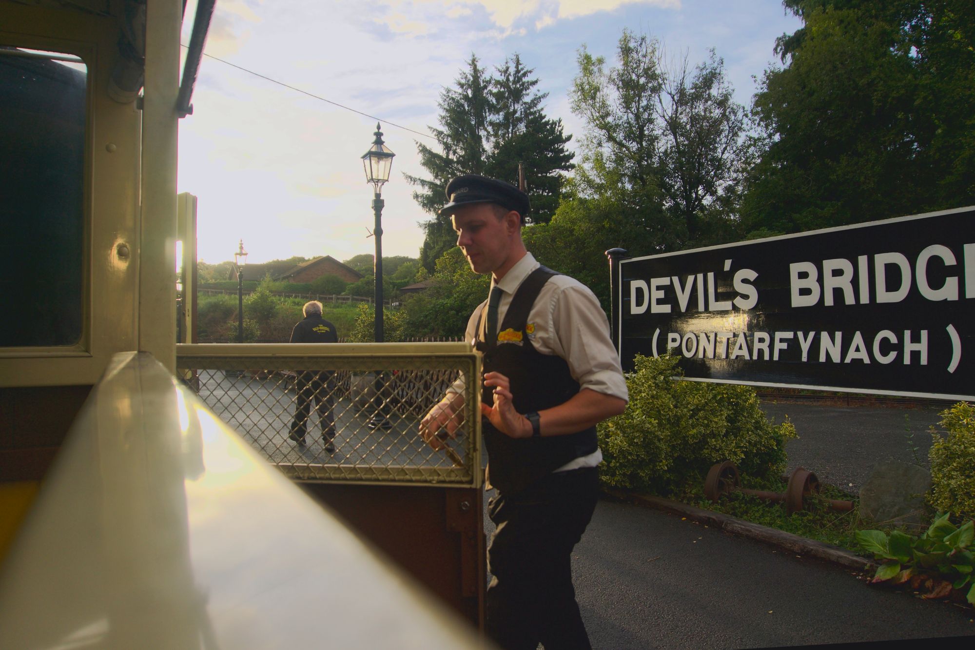 A railway worker in a waistcoat and peaked cap closes a half-hight door to an open railway carriage. The station sign reads DEVIL'S BRIDGE (PONTARFYNACH.)
