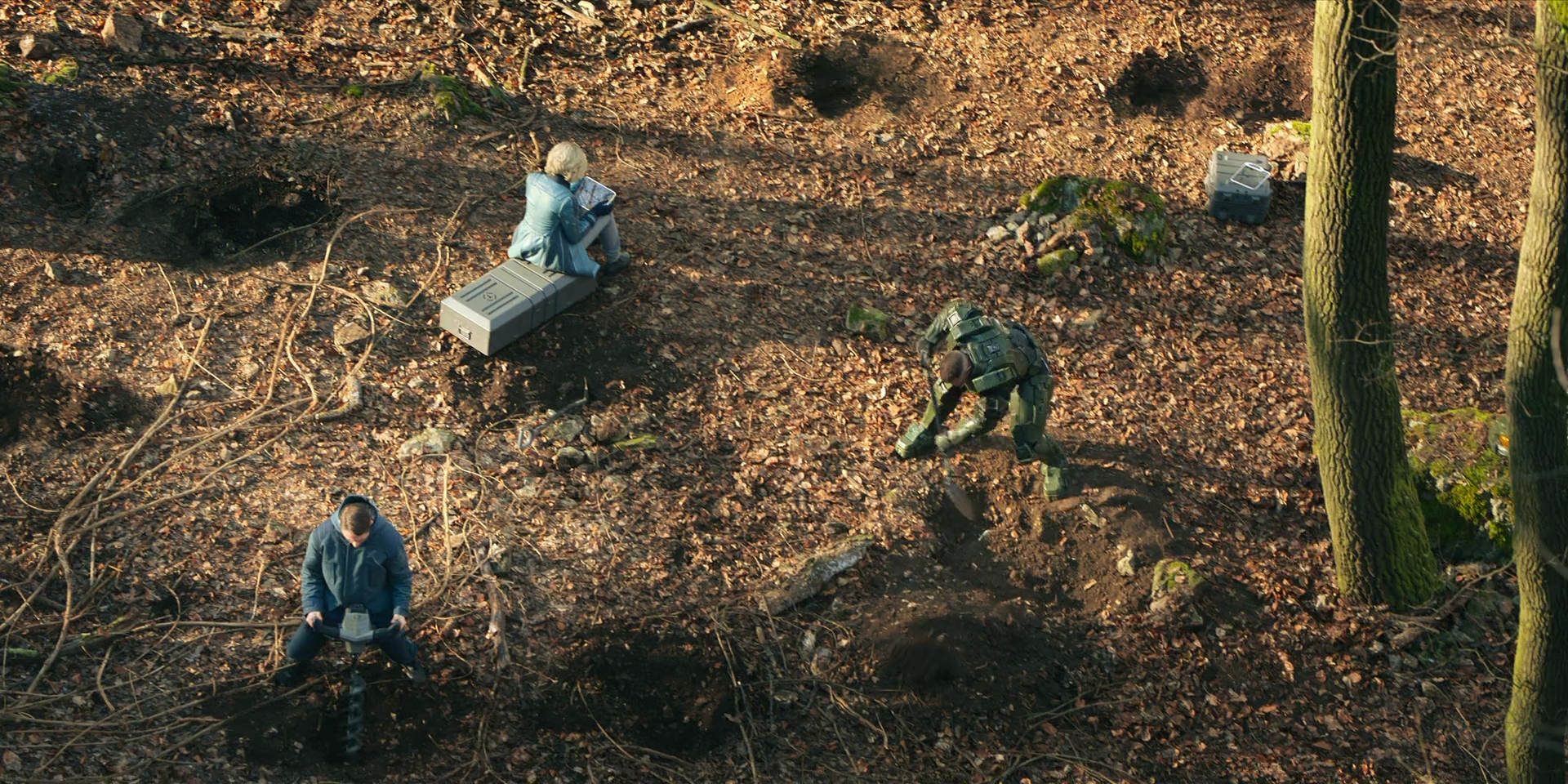 A forest floor. Halsey sits on a crate looking at a tablet computer. Adun squats while using a drilling tool. The Master Chief, with his helmet off, digs with a spade.