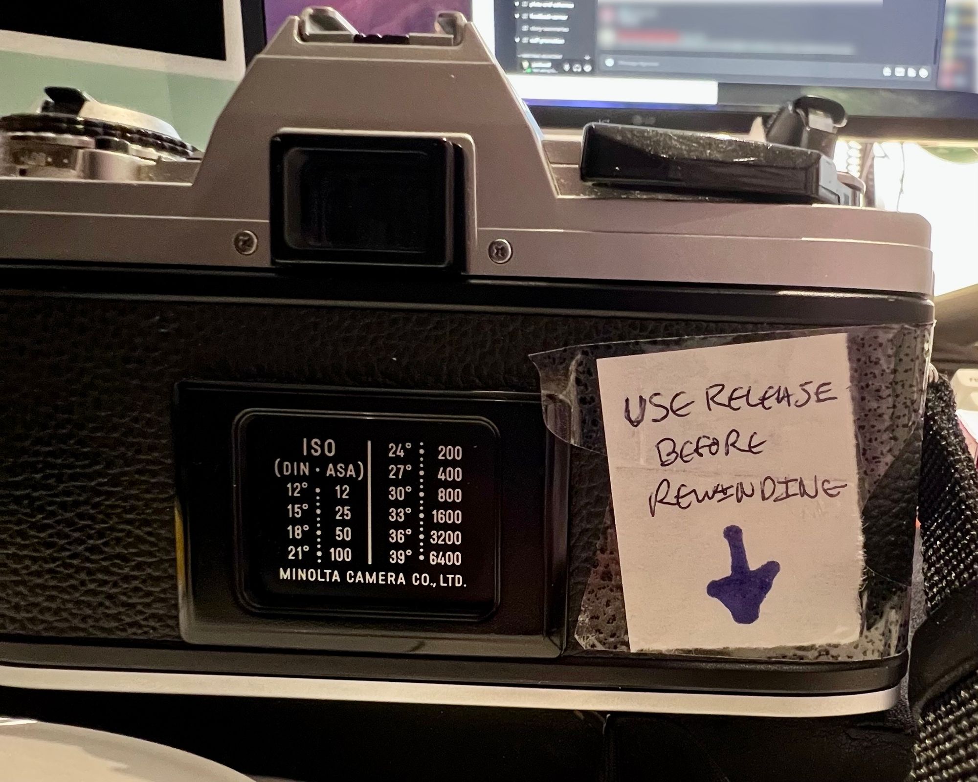 The back of a SLR camera with a note taped to the back saying "USE RELEASE BEFORE REWINDING" and an arrow pointing down.