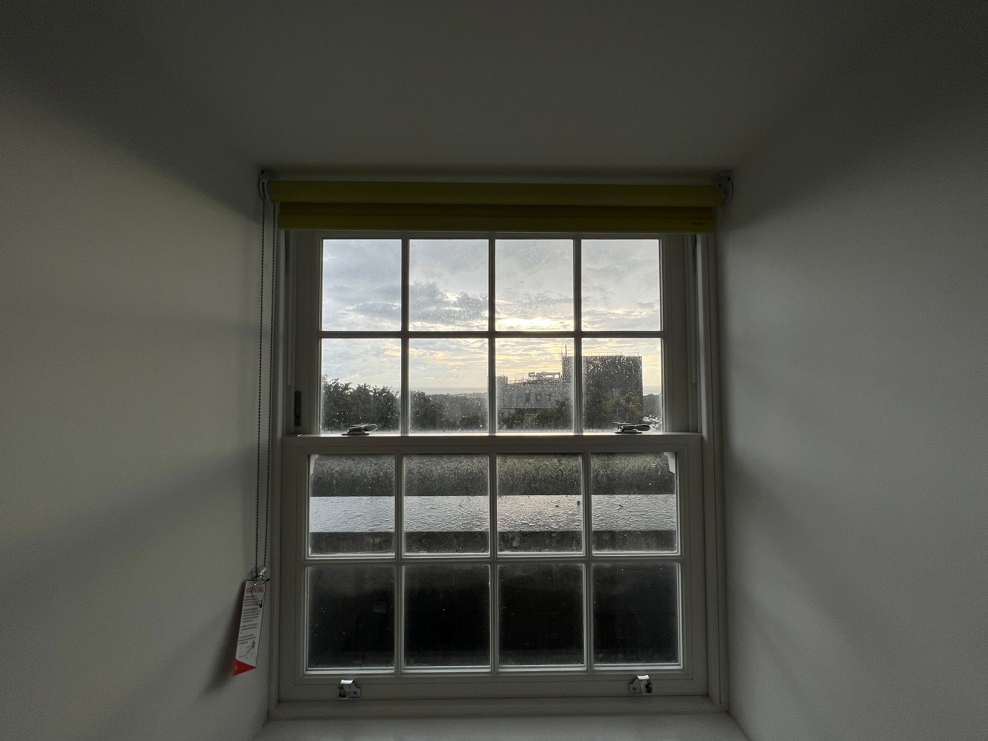 View out of a single glazed window at sunset behind some clouds in the rain. A building and trees are visible, and so is the sea.