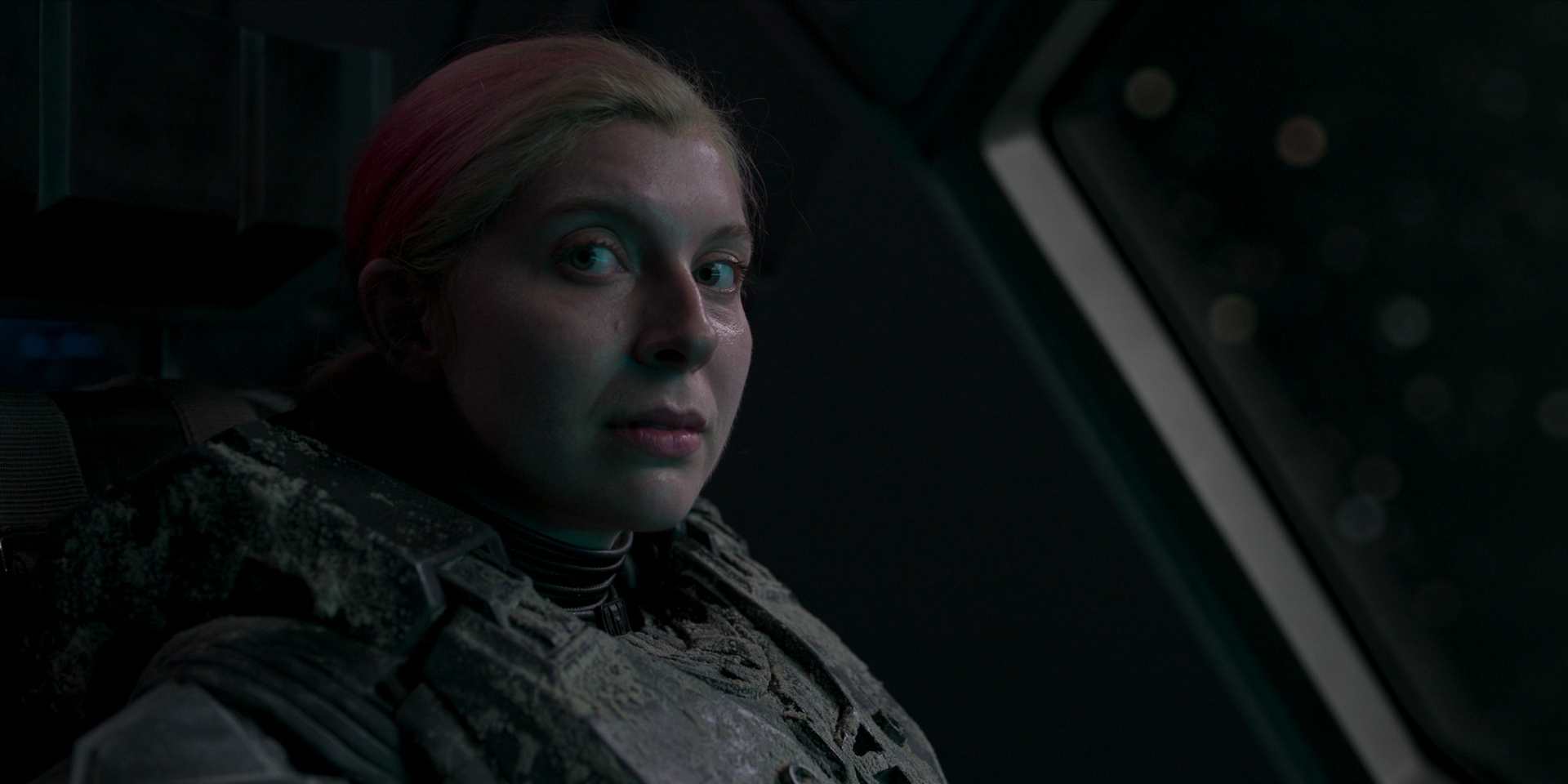 Kai, a white woman with blonde-and-pink hair in a ponytail, sits in a seat looking sideways.