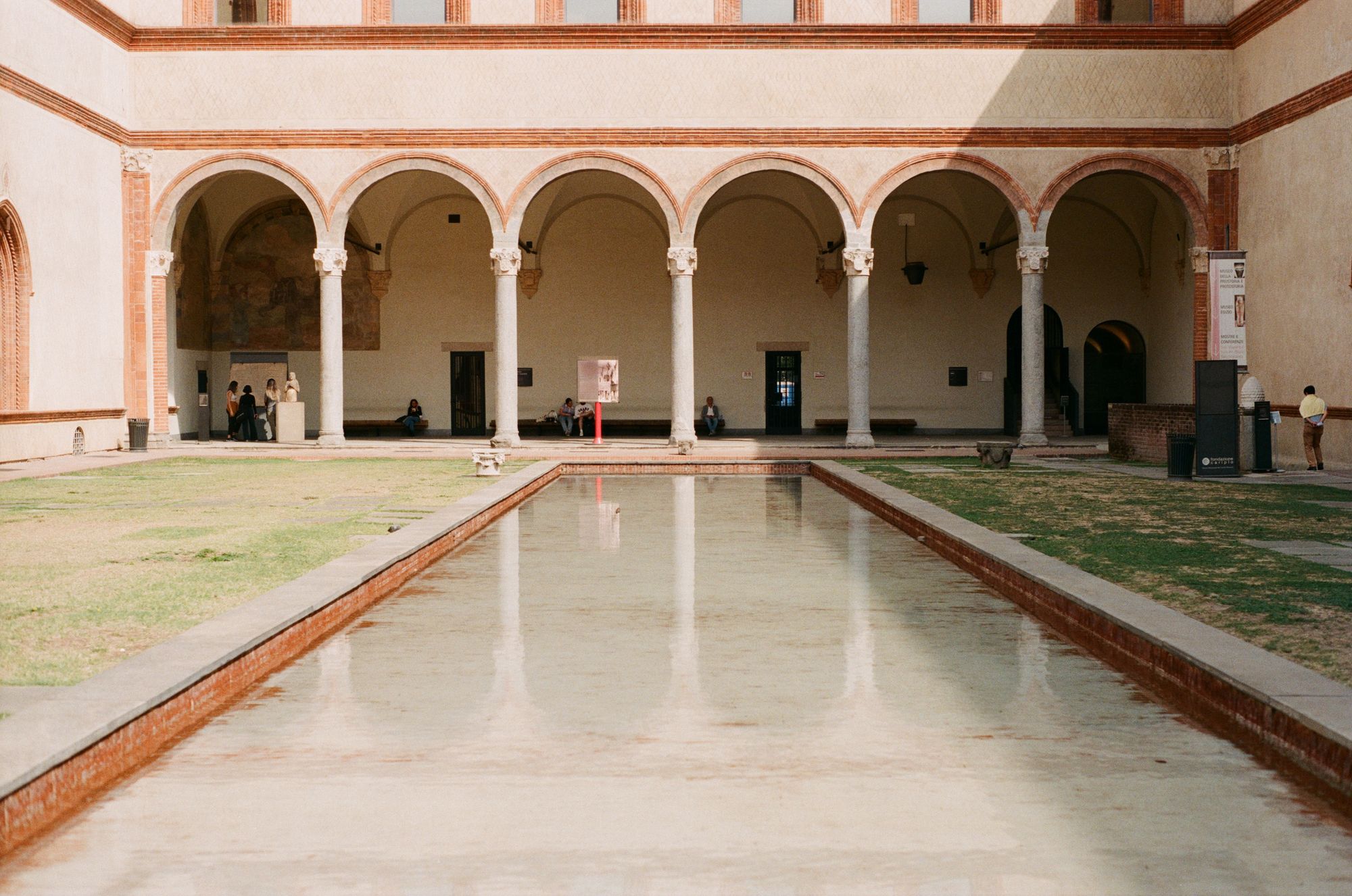 A white colonnade with terracotta detailing, reflecting in a shallow oblong pool