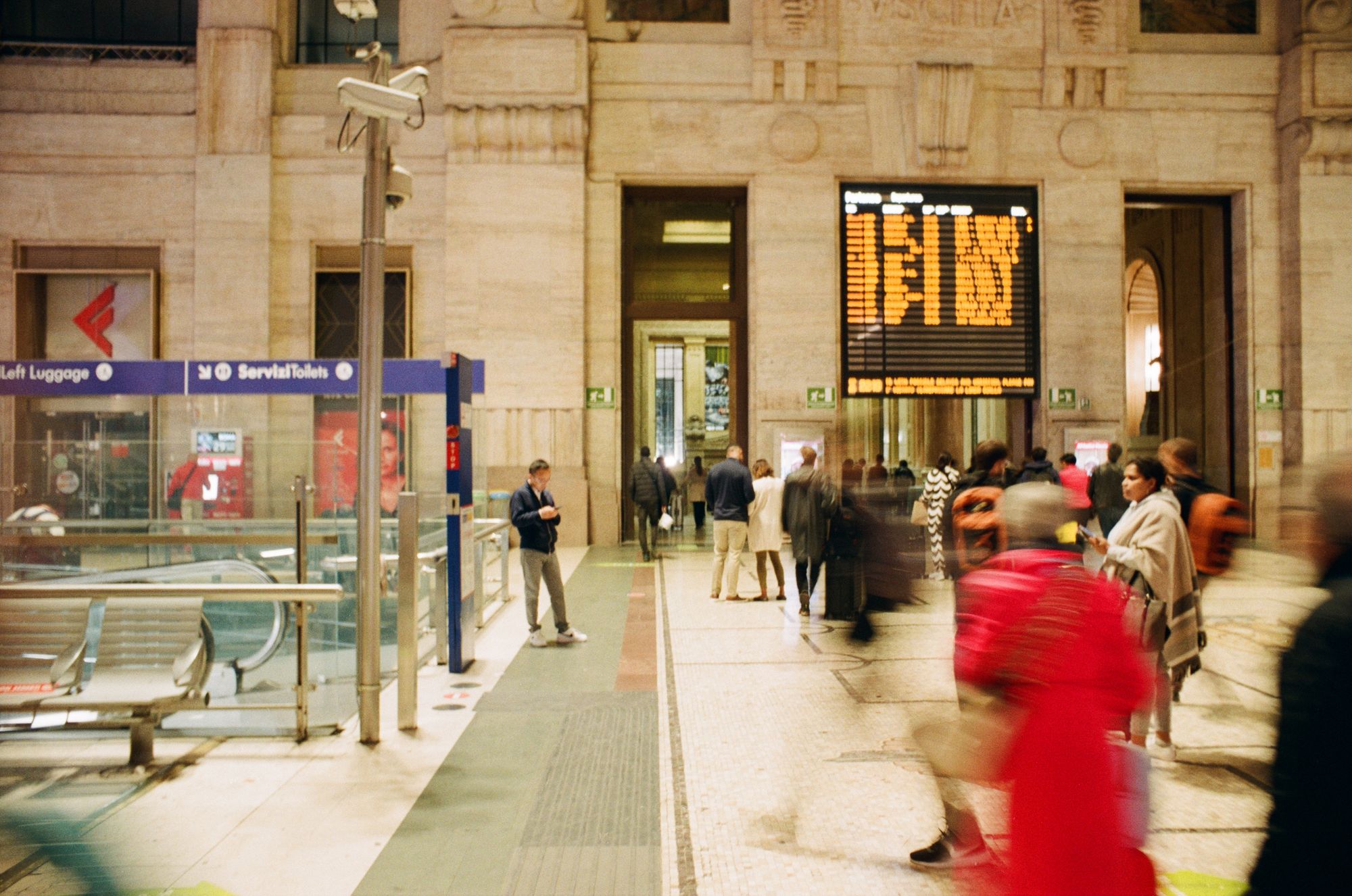 A grainy image of a station concourse with faux marble walls and a departures board, and people bustling around.