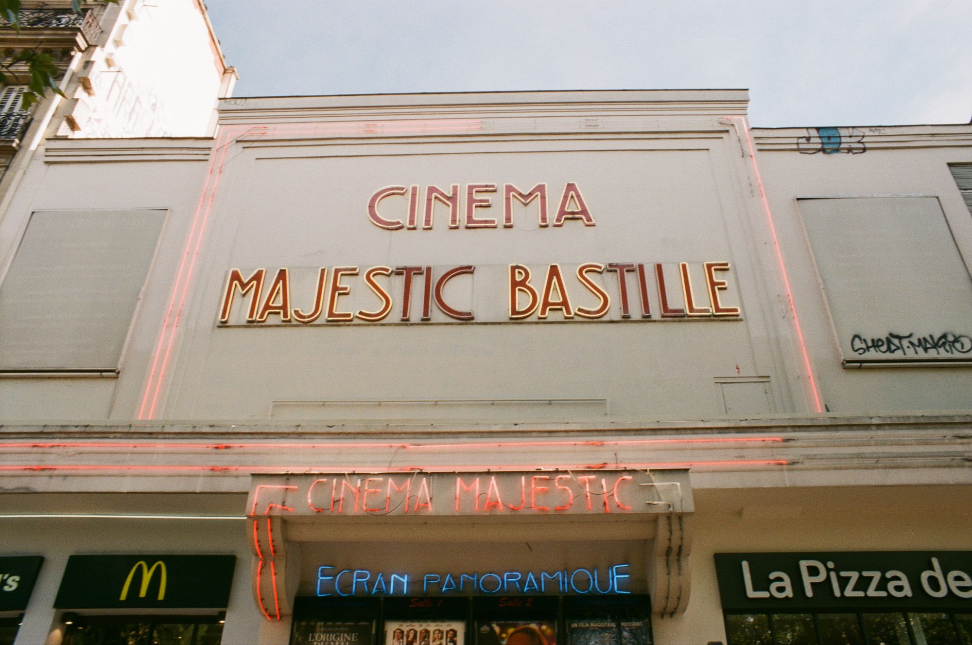 The façade of a large cinema, CINEMA MAJESTIC BASTILLE, with red neon signage and a blue neon strip advertising its ECRAN PANORAMIQUE, with a McDonalds and pizza place on either side.