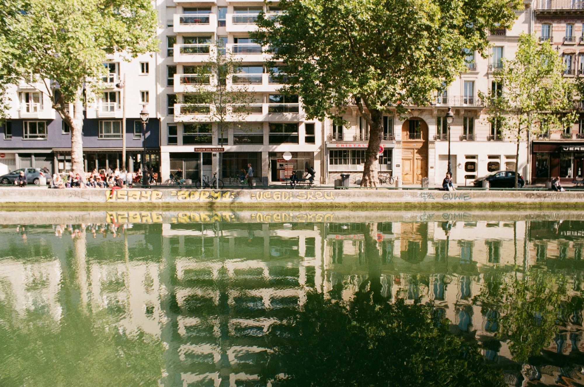 View across a wide canal of a tree-lined Parisian street with graffiti on the canal walls, and people sat by the canal enjoying themselves.